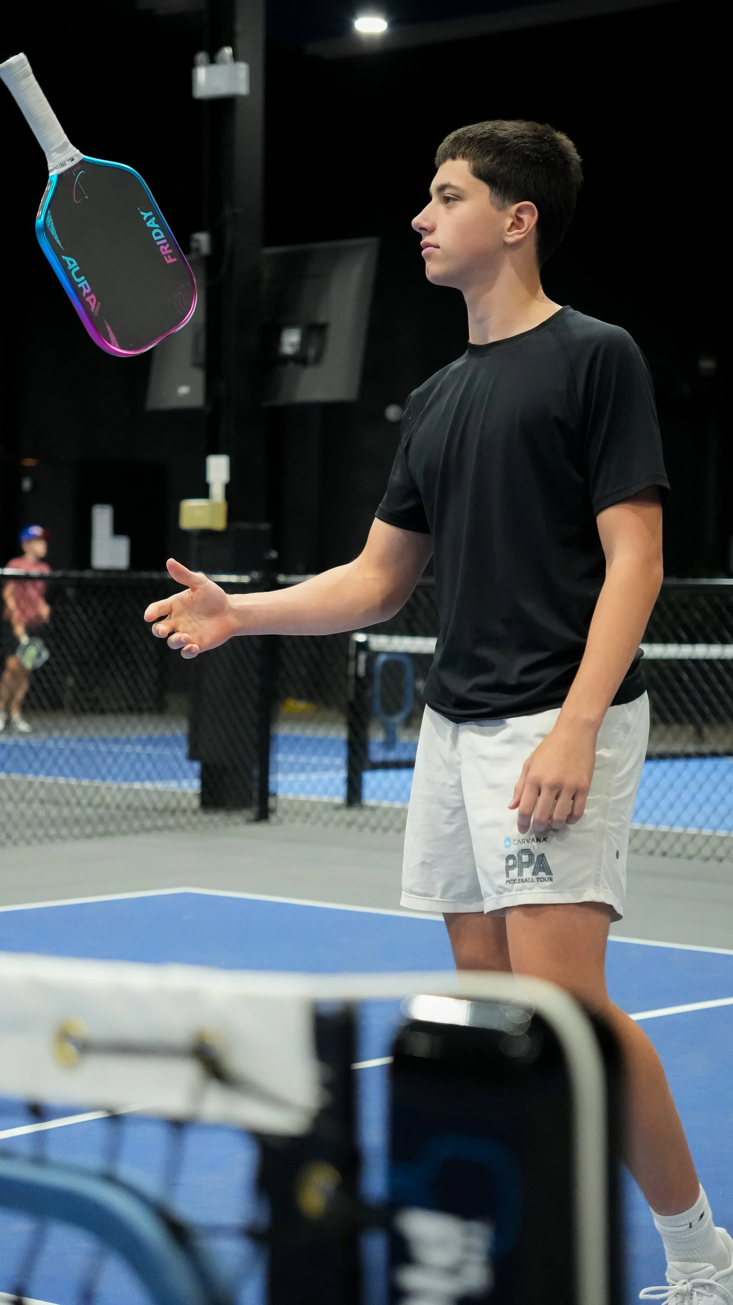 A young male athlete standing on a pickleball court, about to catch a paddle suspended in the air. The court has a blue surface with a chain-link fence, and there are two players in the background, with one wearing a hat.