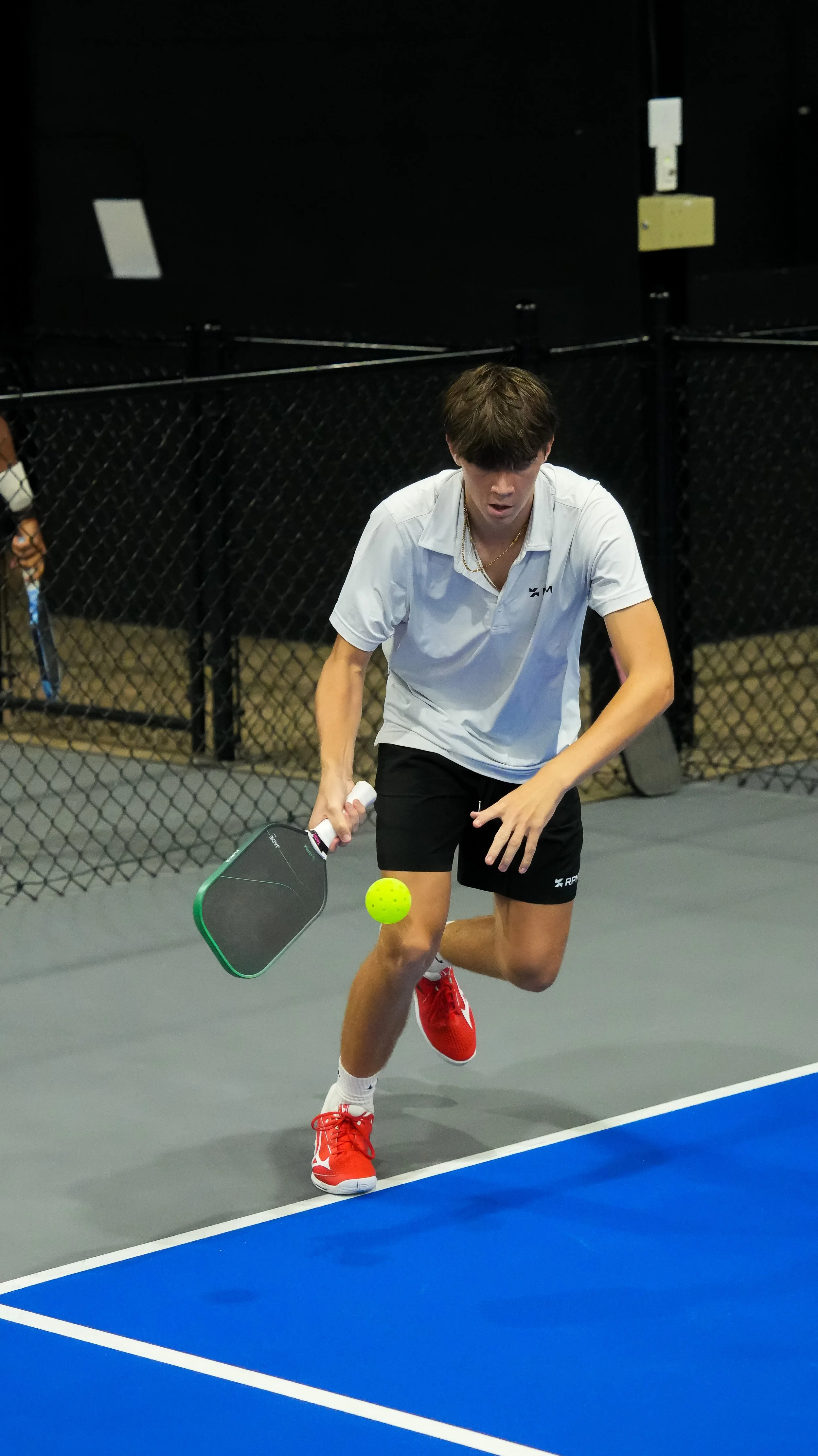 A young man playing pickleball on an indoor court, wearing a white shirt, black shorts, and red shoes, holding a paddle near a yellow pickleball.