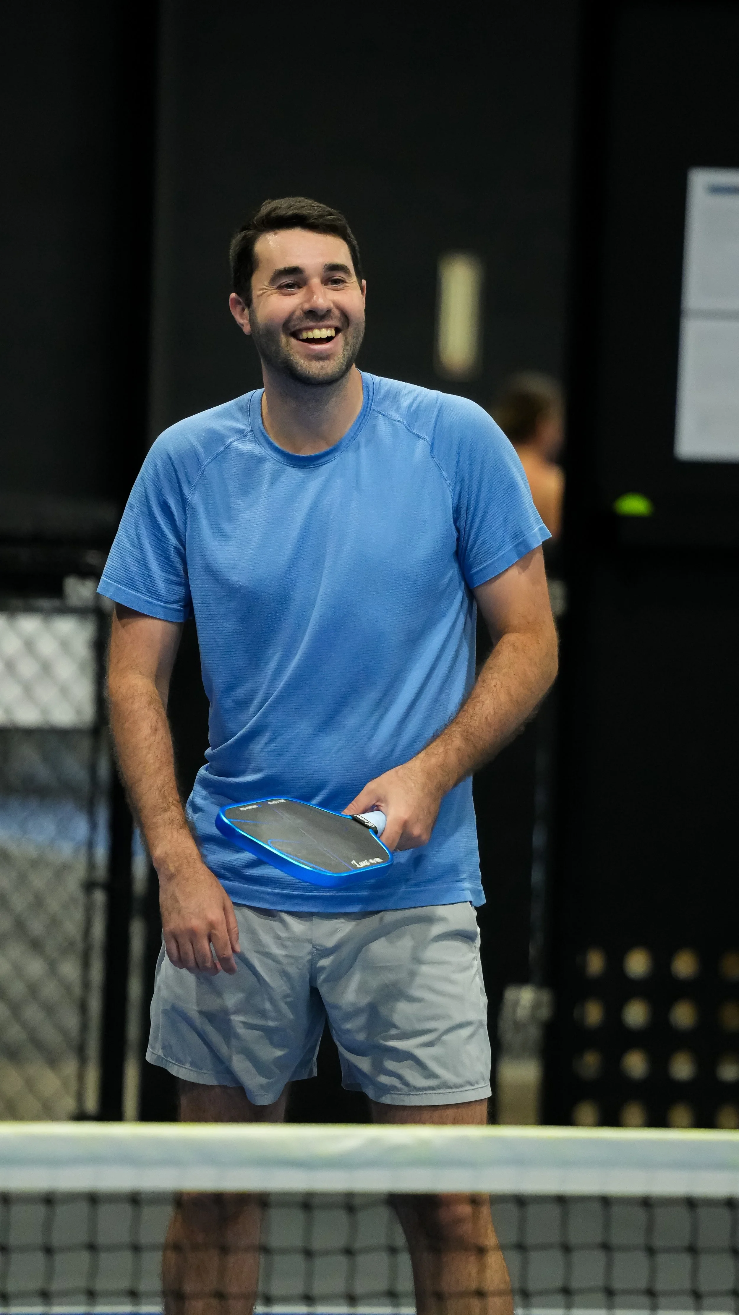 A man smiling and holding a tennis racquet wearing a blue shirt and light shorts on a tennis court.