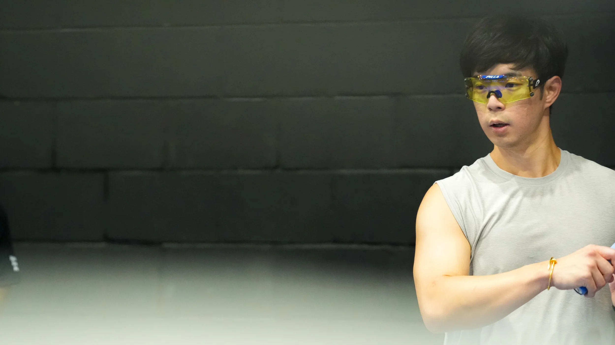 Young man wearing yellow safety glasses and a sleeveless gray shirt, standing against a dark wall, looking to the side.