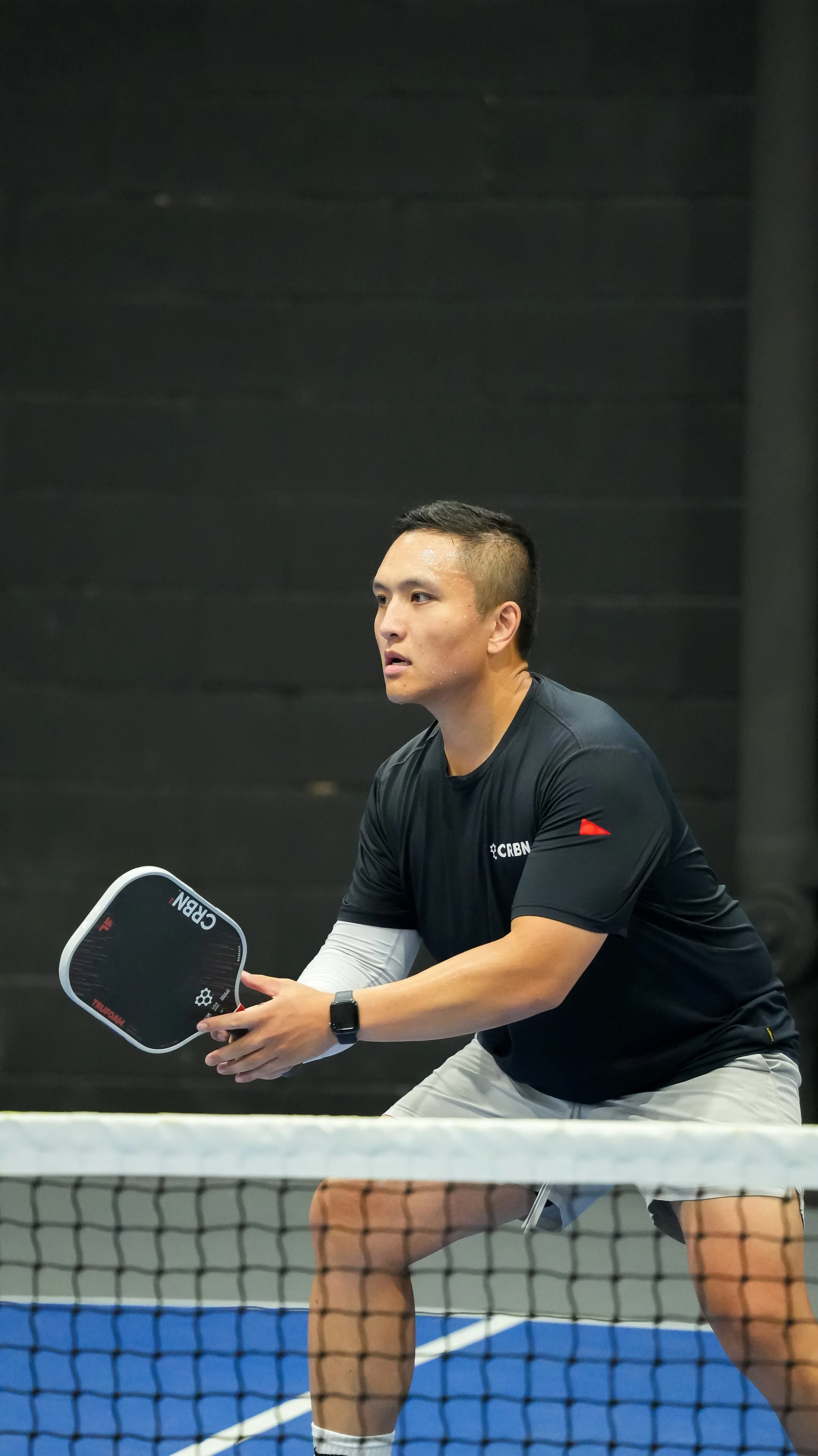 A man playing pickleball, holding a paddle, on an indoor court with a net in front of him.