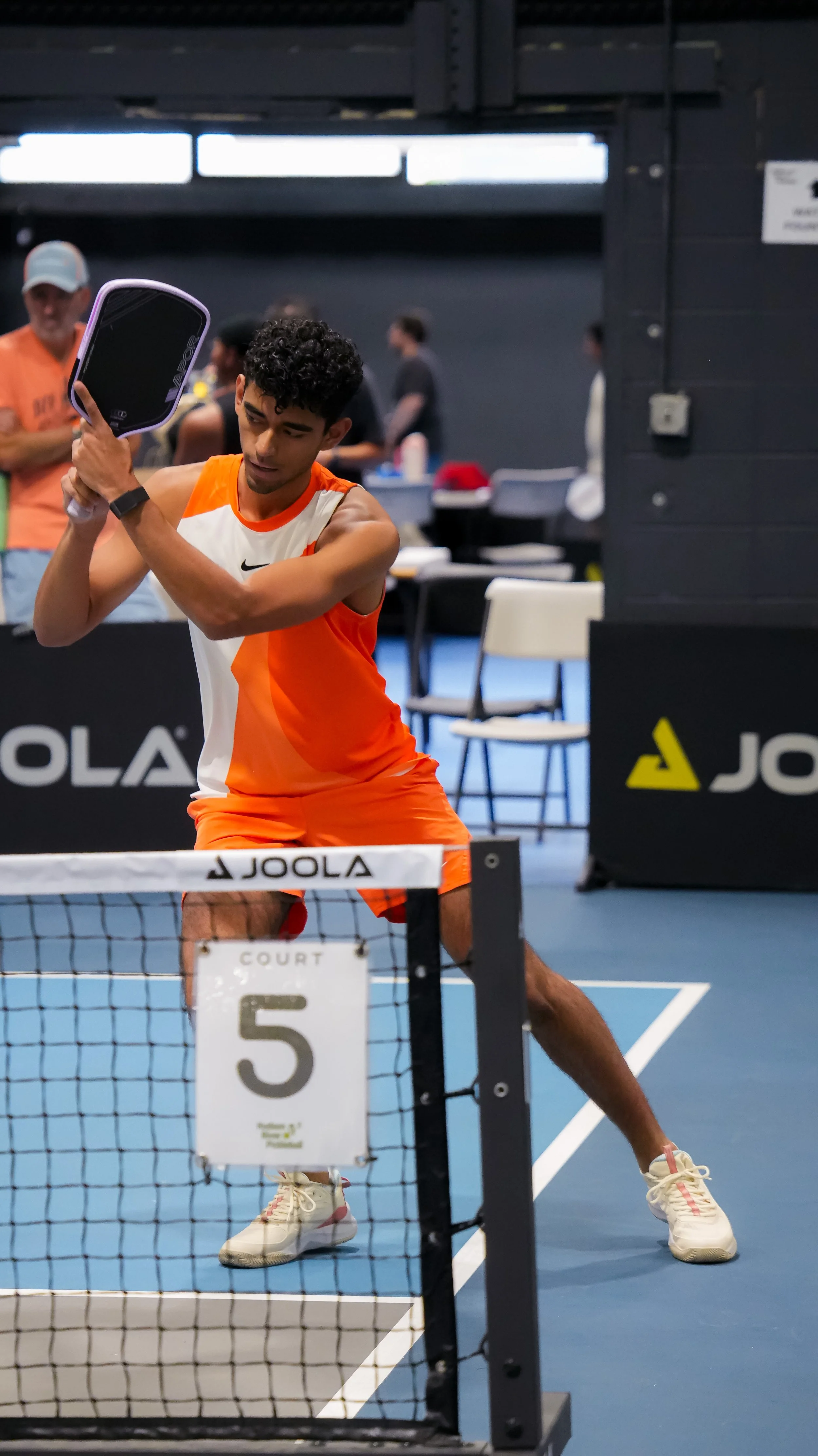 A male athlete in an orange and white sports uniform playing table tennis in an indoor sports facility, with spectators and other players in the background.