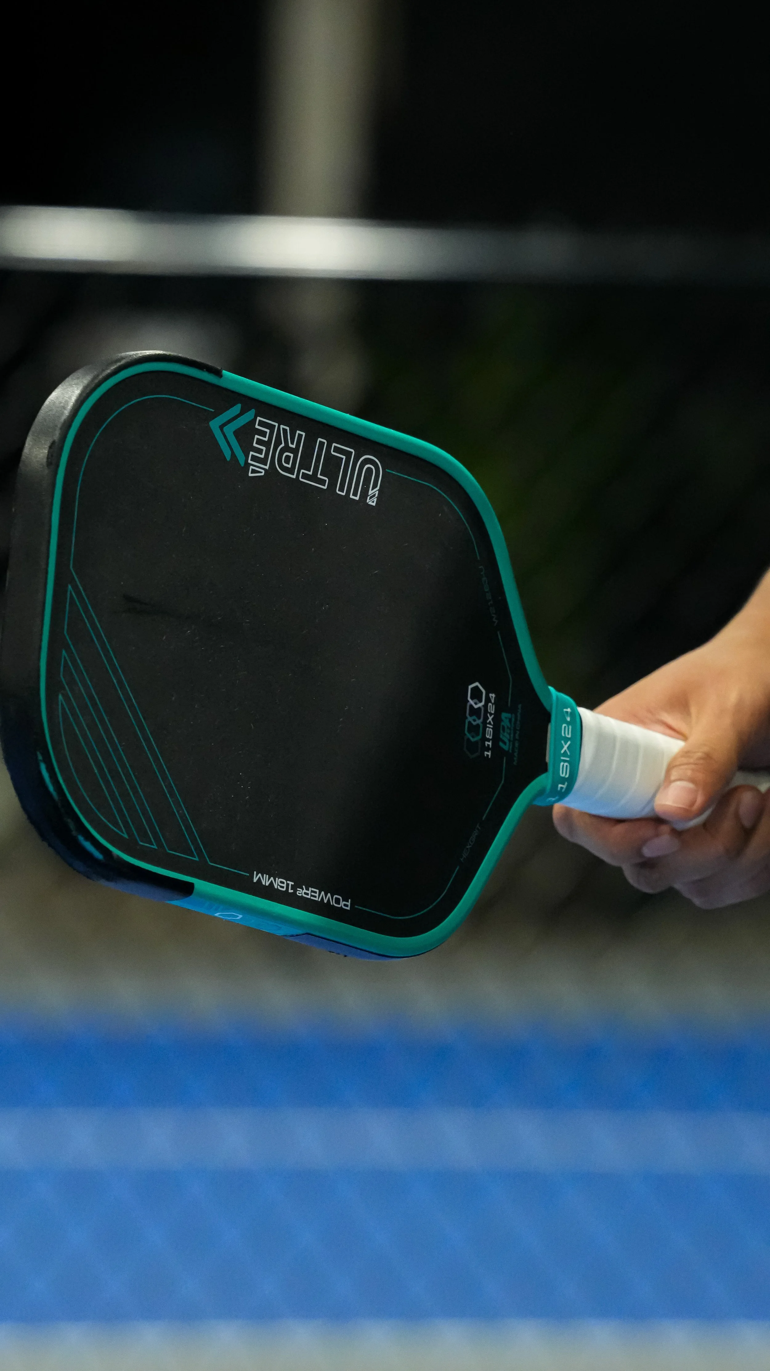 Person holding a black and teal PowerGear 16MM paddle tennis racket with their hand visible, with a blurred background.