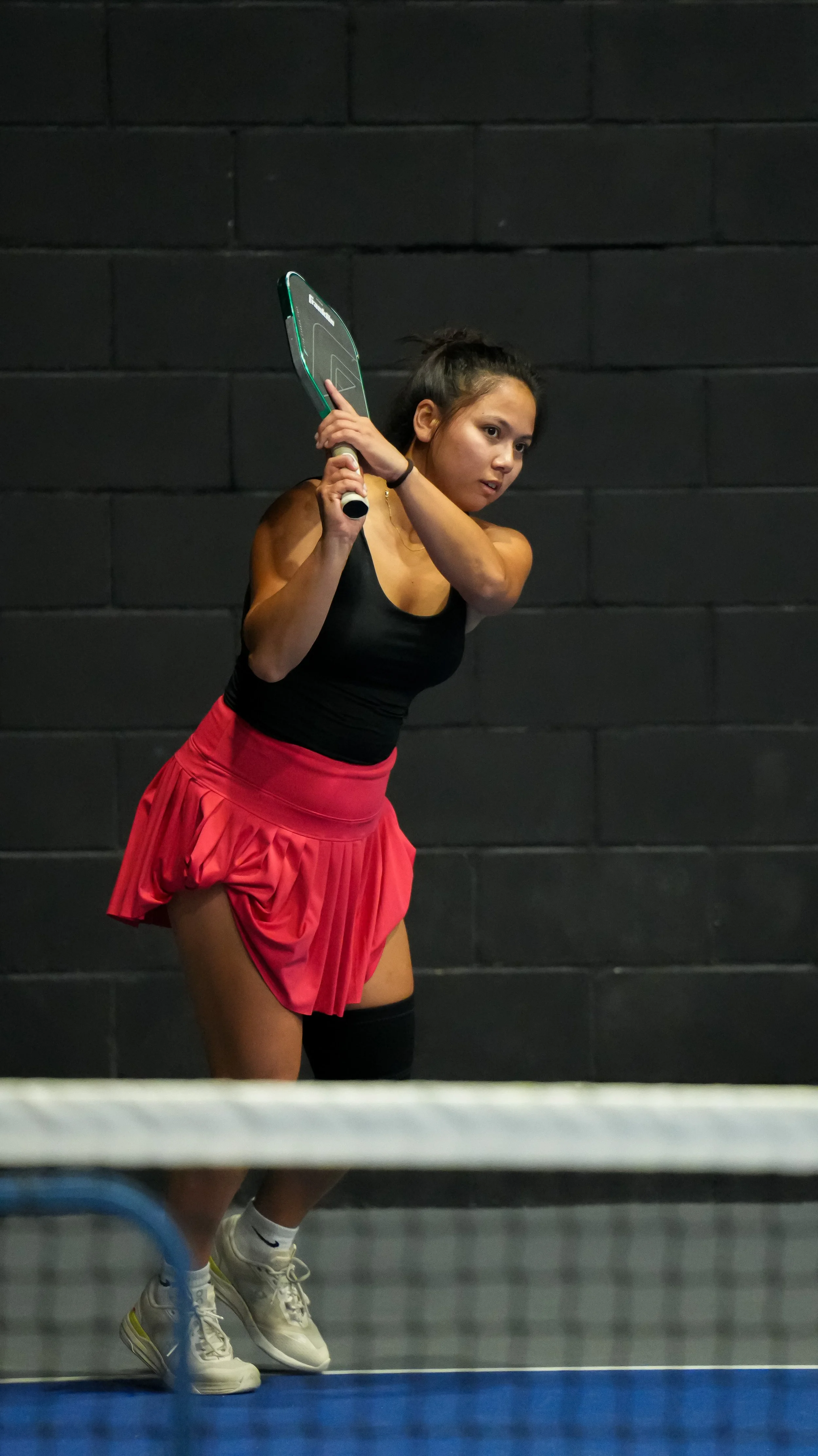 A woman playing tennis indoors, wearing a black tank top, red skirt, and white tennis shoes, holding a tennis racket ready to hit a ball.