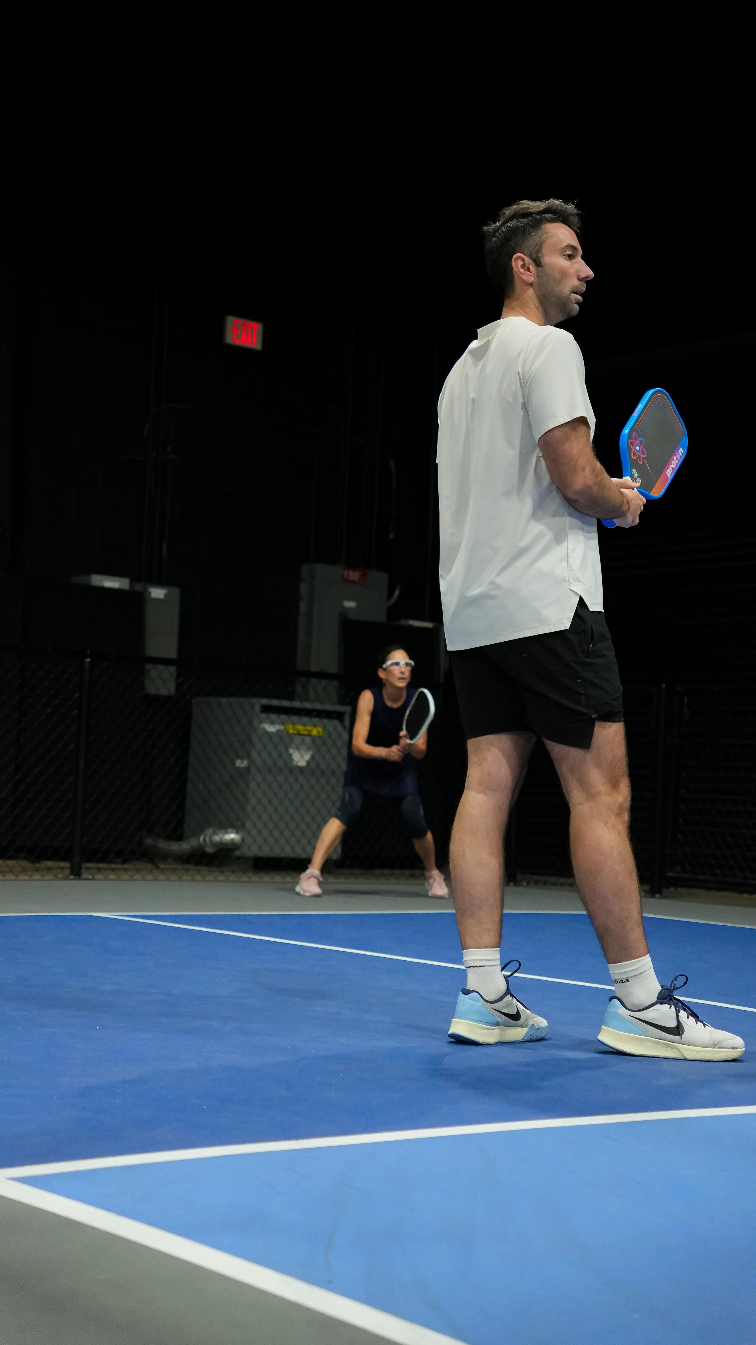Two people playing pickleball on an indoor court, one man standing in foreground holding a paddle, woman in background ready to hit the ball. Black walls with red exit signs.