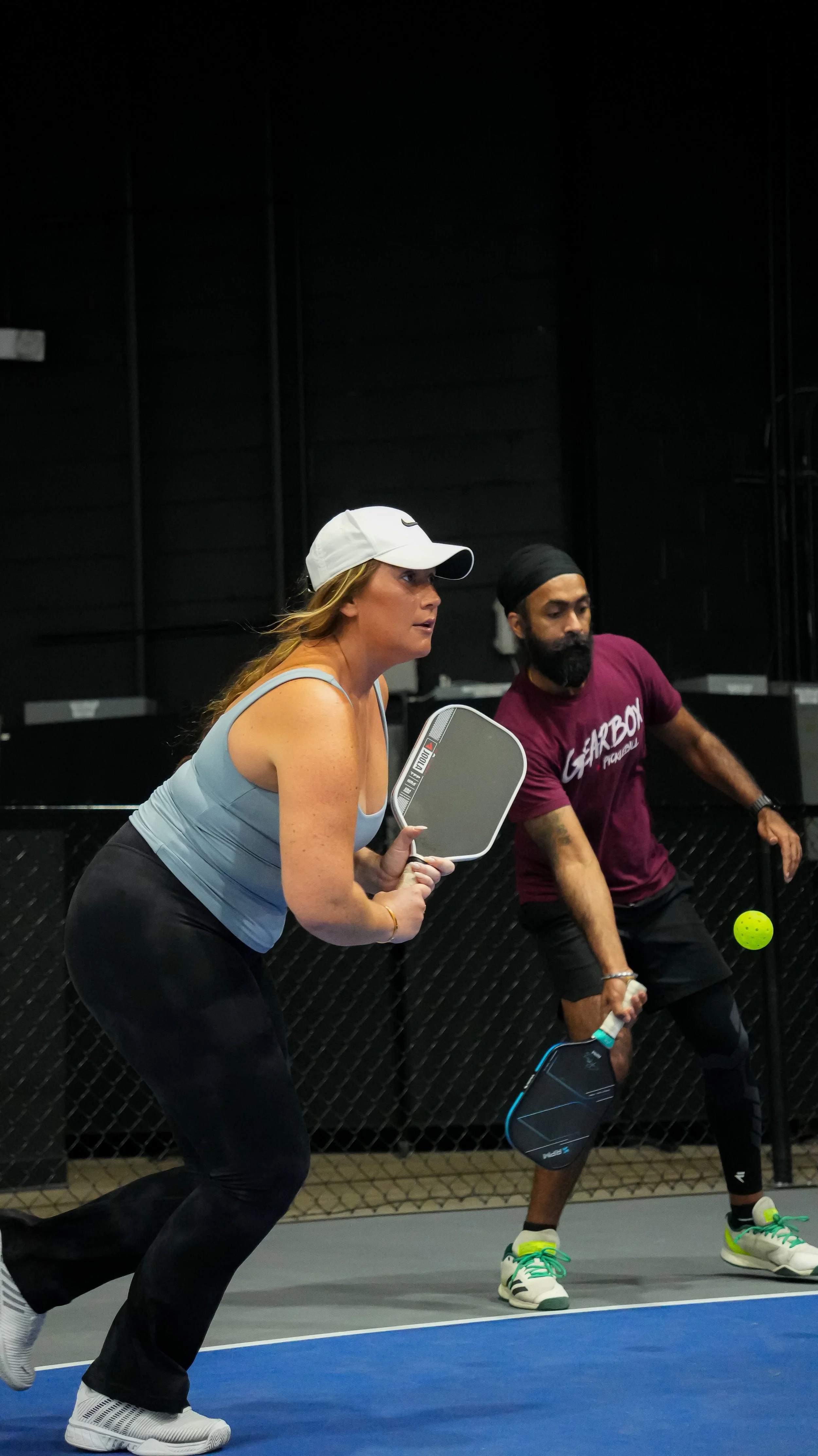Woman and man playing pickleball on indoor court, woman holding paddle and preparing to hit yellow ball.