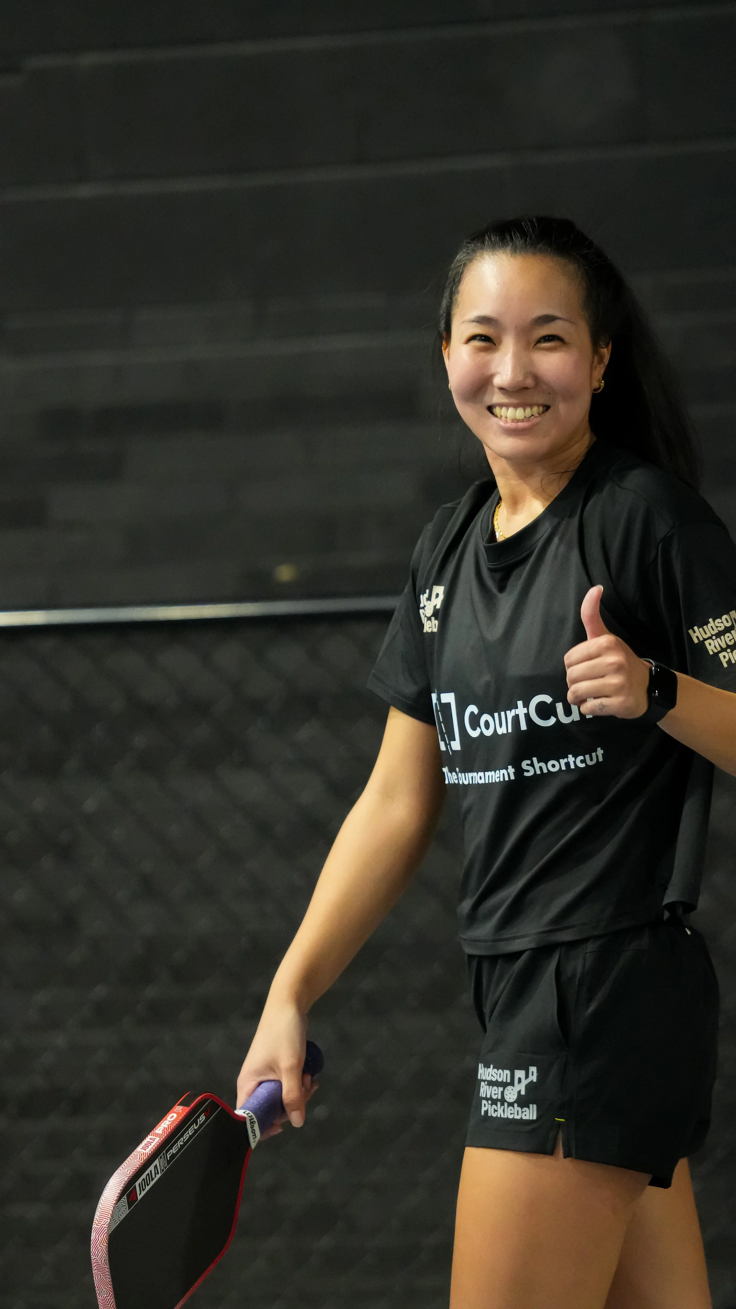 A woman smiling and giving a thumbs-up while holding a paddle, dressed in black sportswear with 'CourtCure' and 'Hudson River Pickleball' printed on it, in an indoor sports facility with a dark wall in the background.