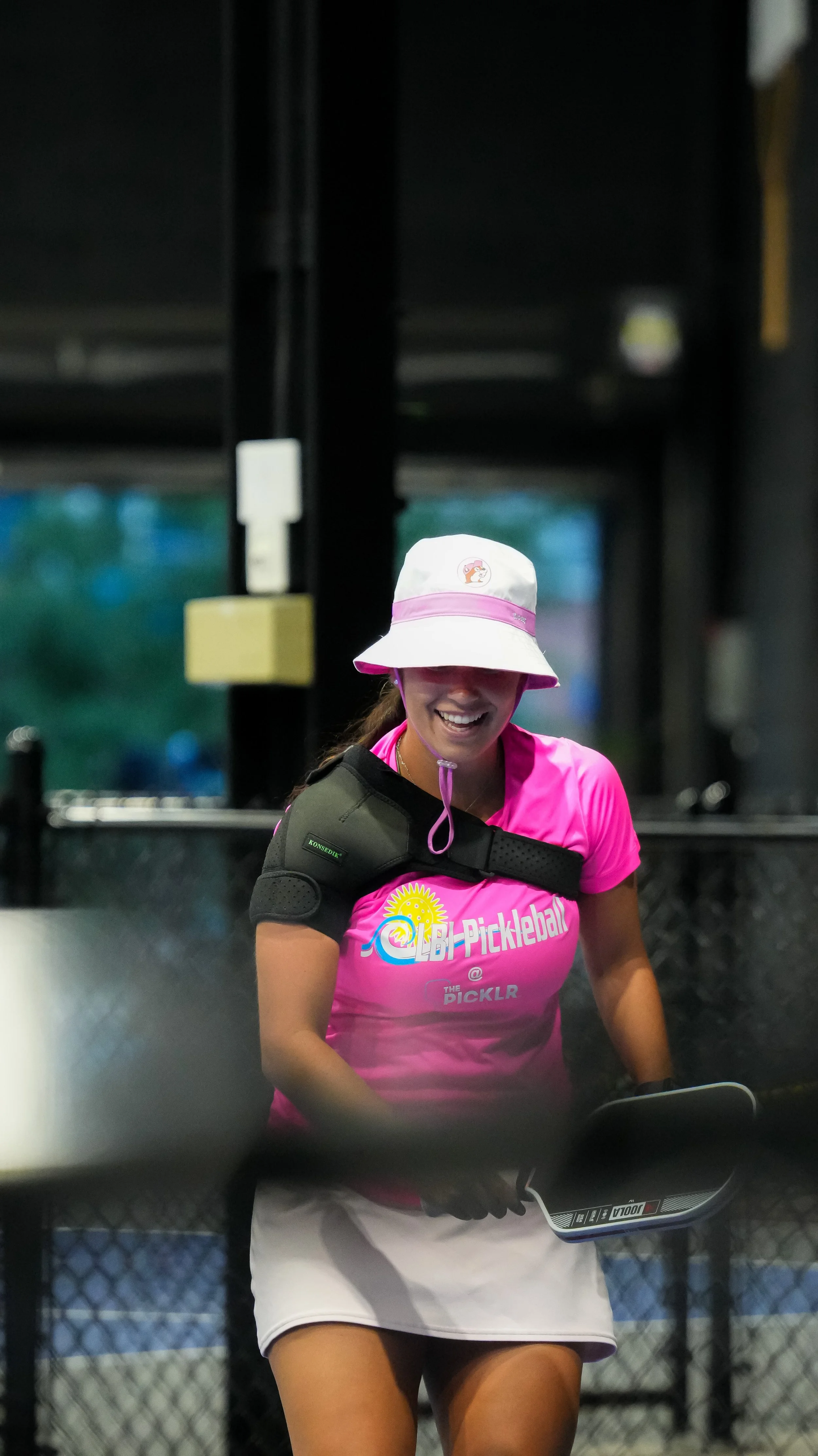 A woman smiling, wearing a pink sports shirt with a logo, a white hat with a logo, and a shoulder brace, standing behind a fence with a blurred outdoor background.