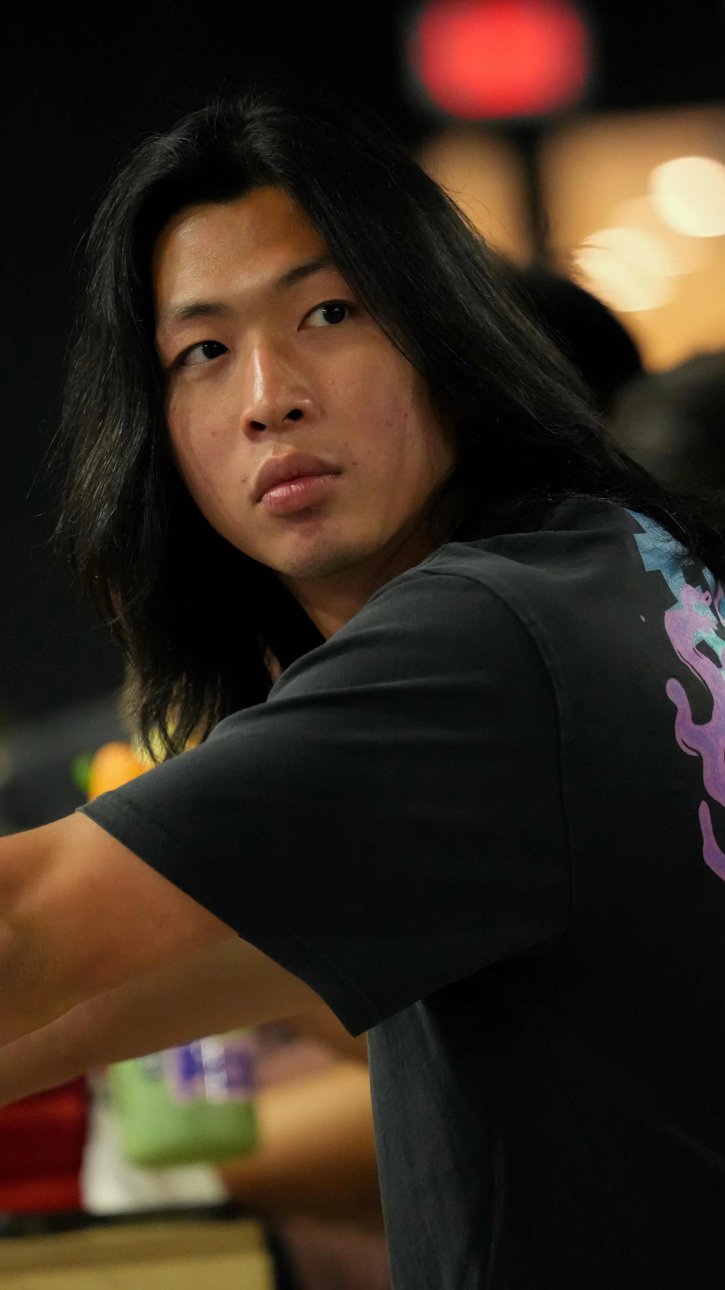 A young person with long black hair and a serious expression, wearing a black T-shirt, sitting at a table in a dimly lit environment with blurred background lights.