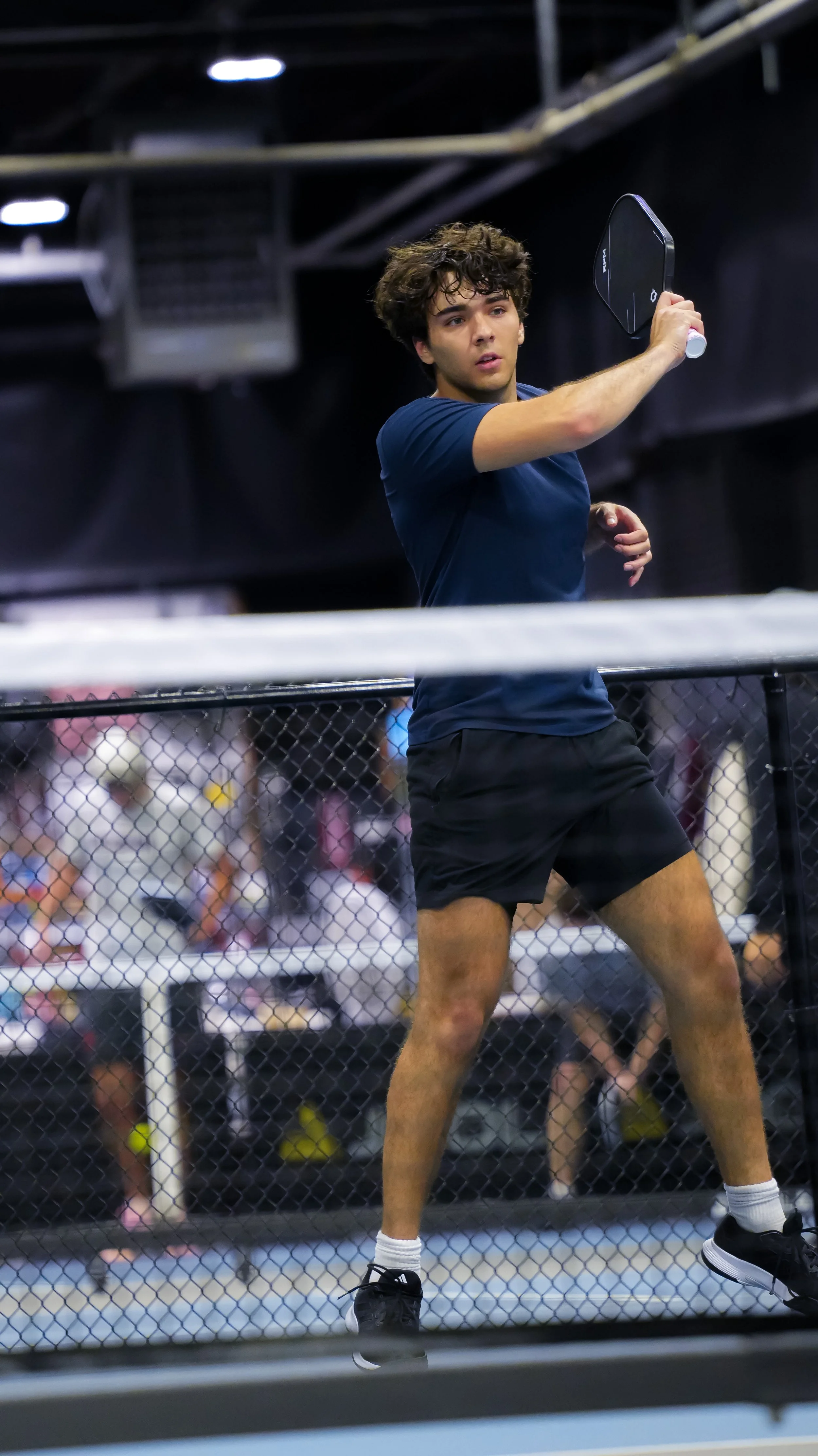 Young man playing pickleball inside an indoor sports facility, holding a paddle in one hand and standing near a net.
