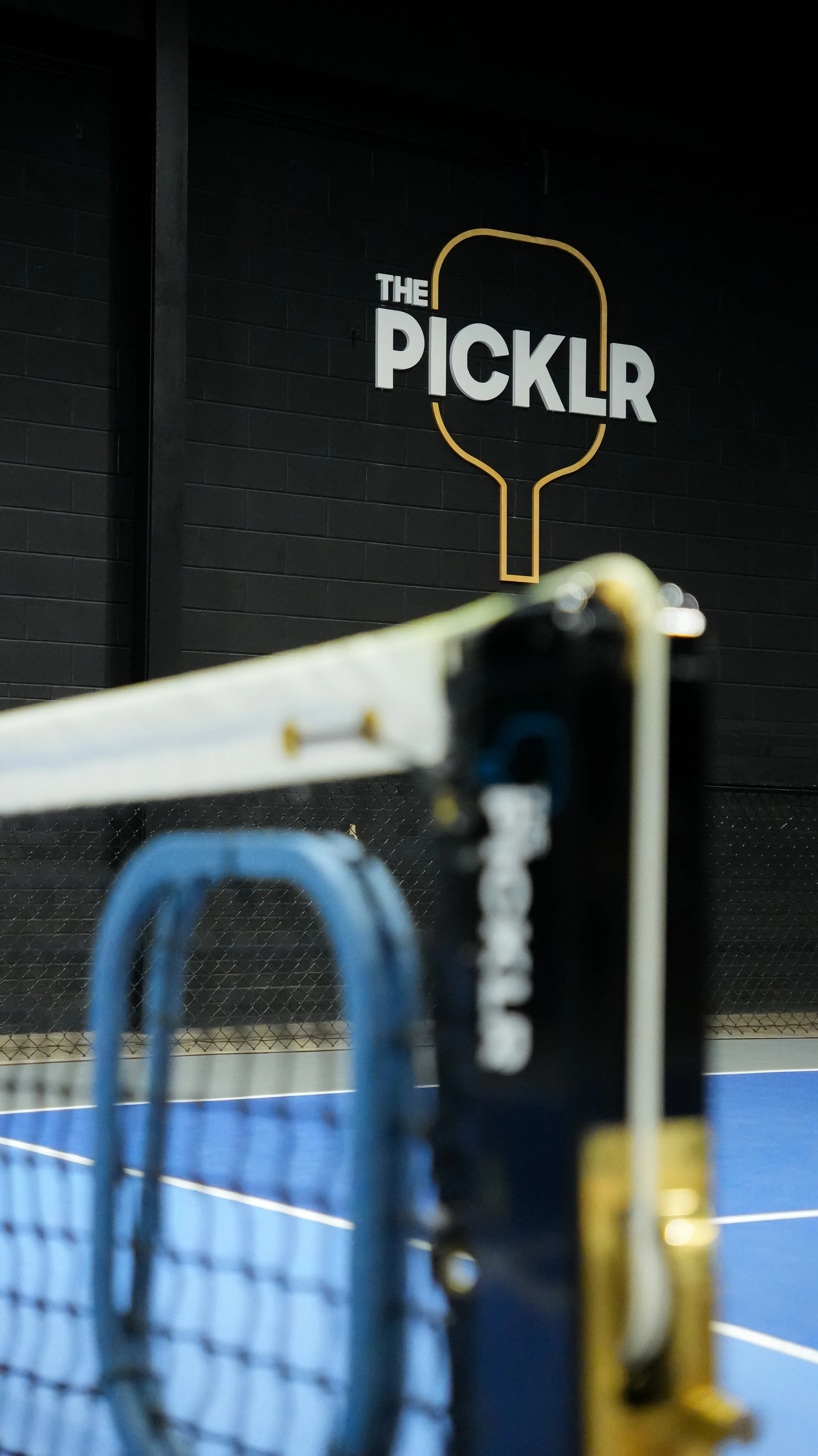 View of a sports facility with a sign that reads 'The PICKLR' on a black brick wall, with the foreground showing the top of a tennis or badminton net and some blue and black sports equipment.