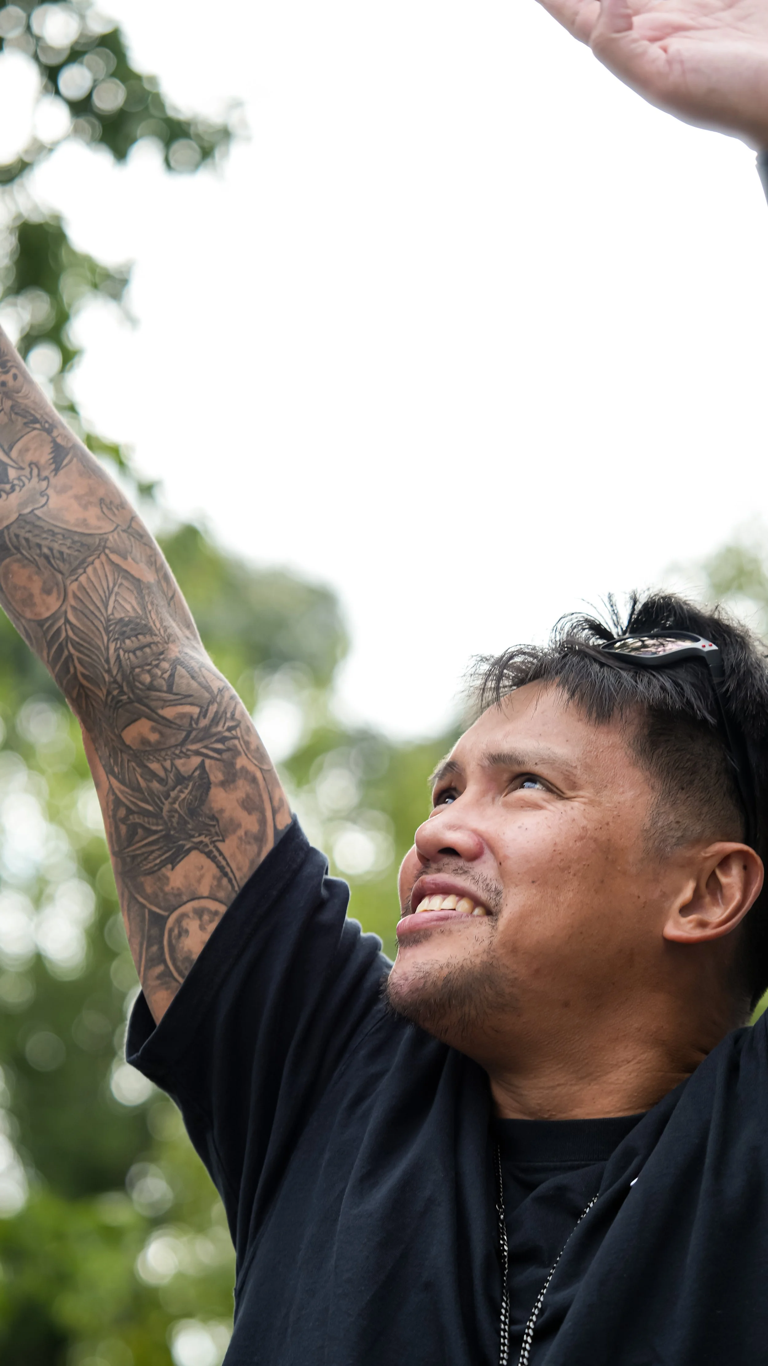 A young man with a tattooed arm smiling and looking upwards, wearing sunglasses on his head and a black shirt, outdoors with blurred green trees in the background.