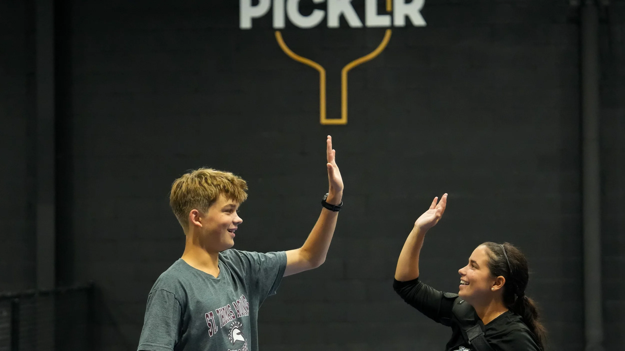 A young man and woman with dark hair high-five each other indoors, with a black wall and a sign that says 'PICKLE' above them.