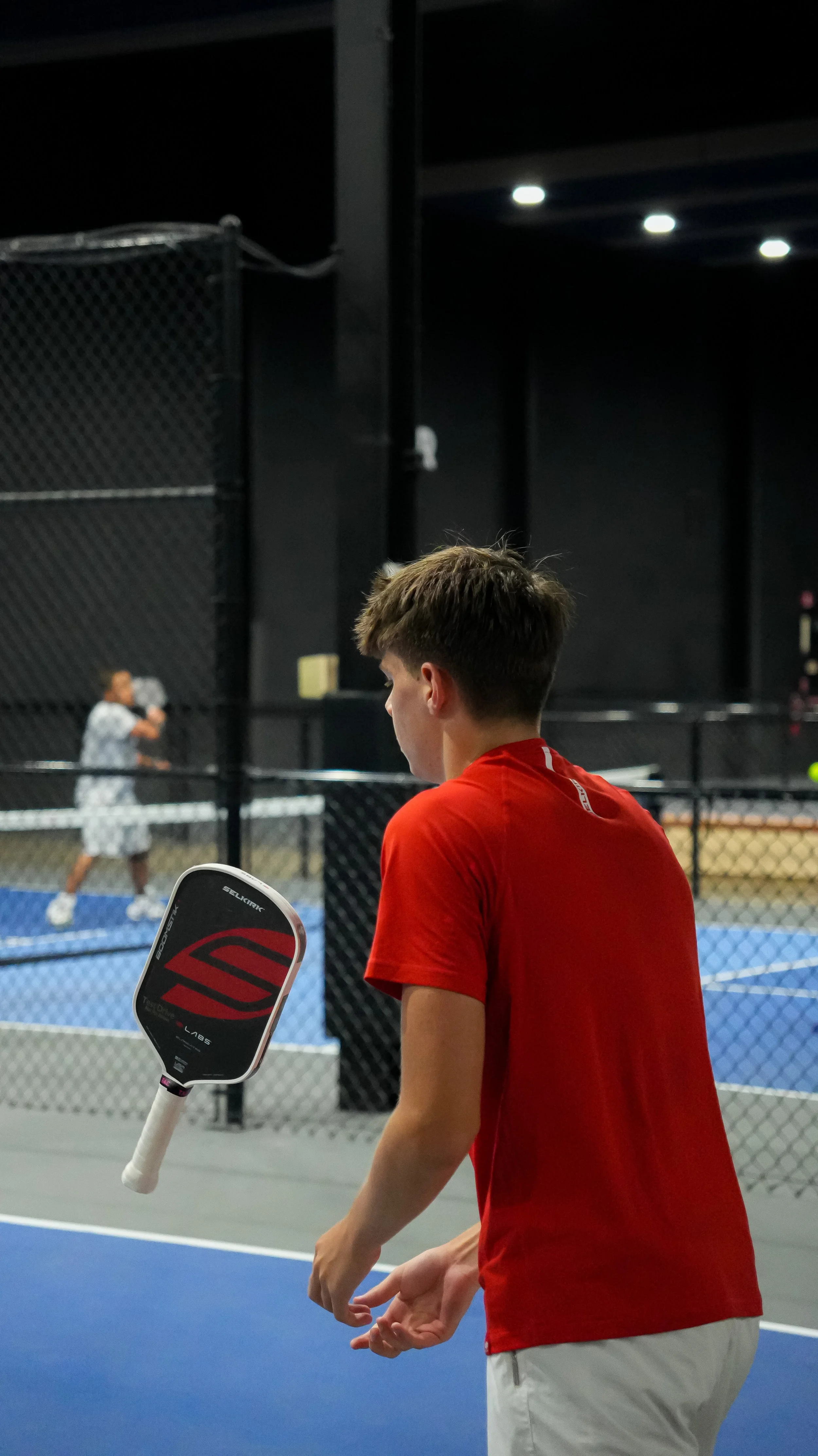 Young man in red shirt holding a padel racket, standing in an indoor padel court, with a woman playing in the background.