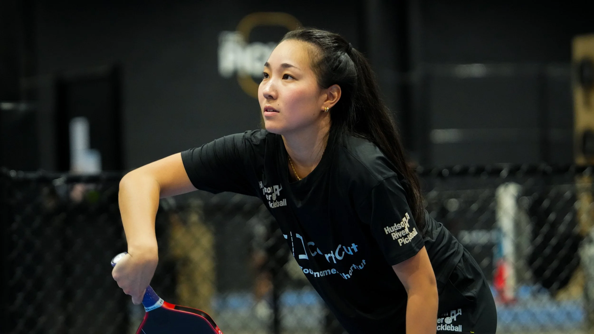 A woman with long dark hair in a braid, wearing a black T-shirt with white text, playing pickleball indoors, holding a paddle and ball, with a black gym background.