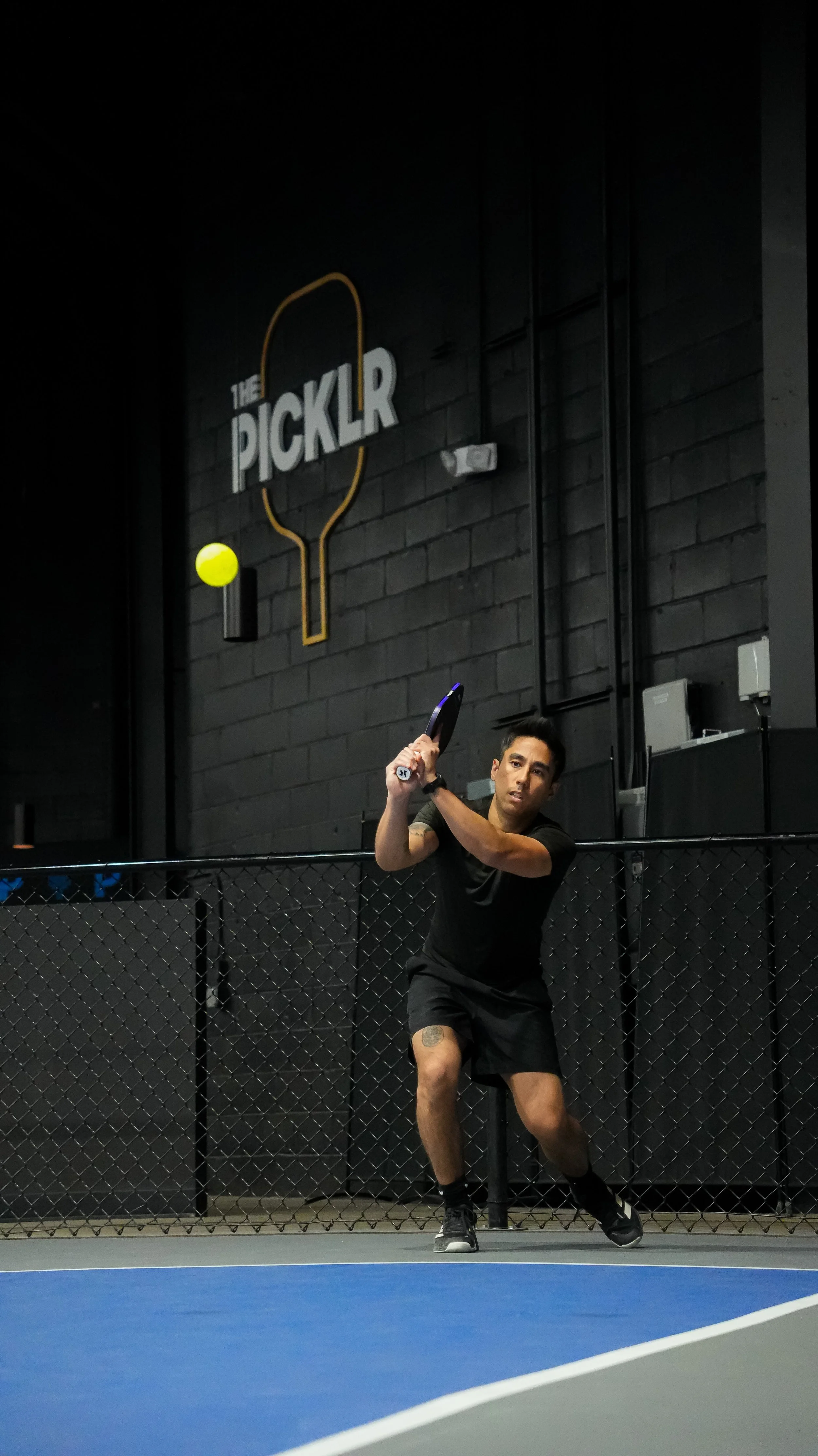 A man playing pickleball on an indoor court, holding a paddle, with a black wall and the Pickleball logo in the background.
