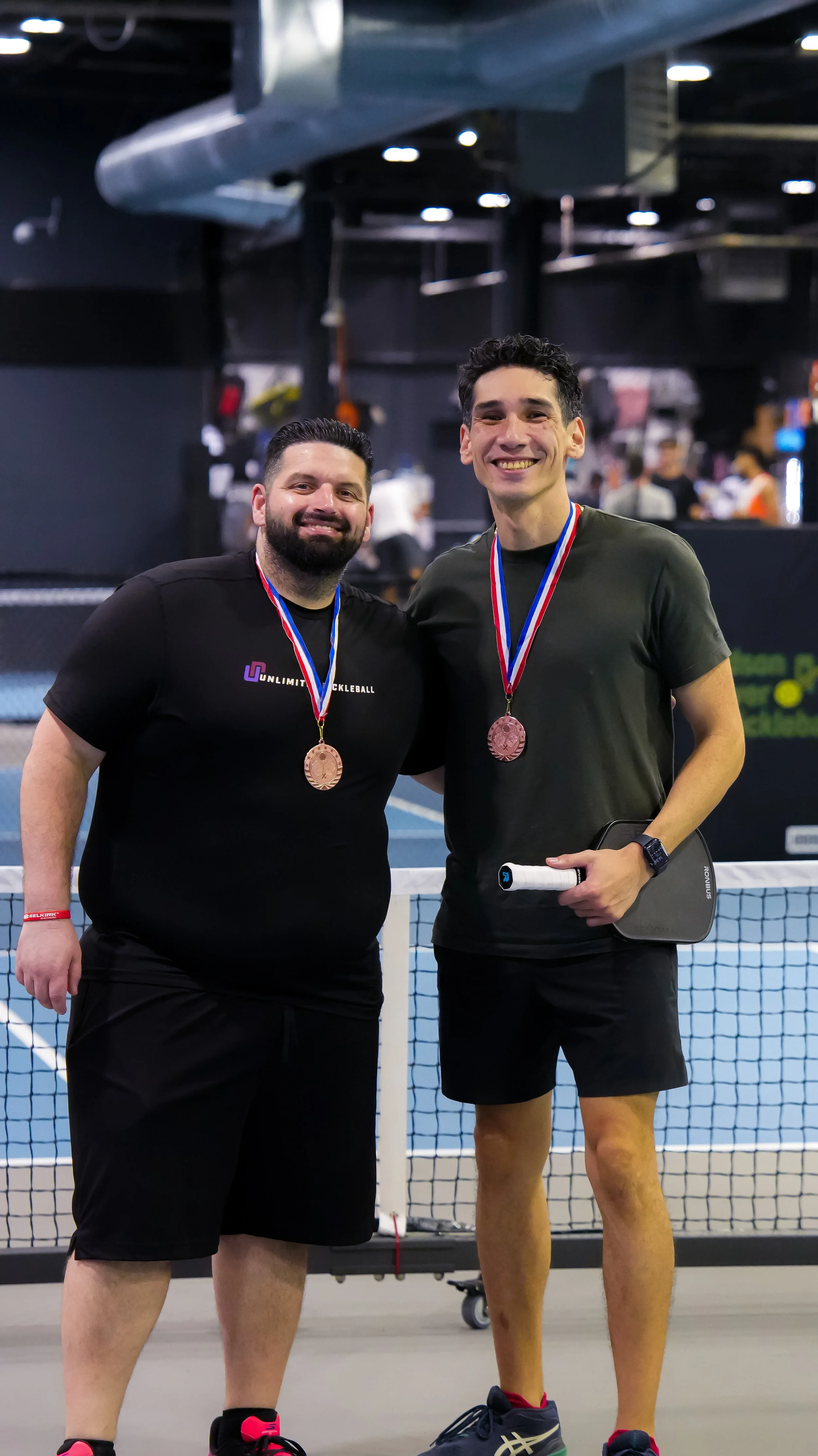 Two male tennis players smiling and wearing medals around their necks, standing on an indoor tennis court.