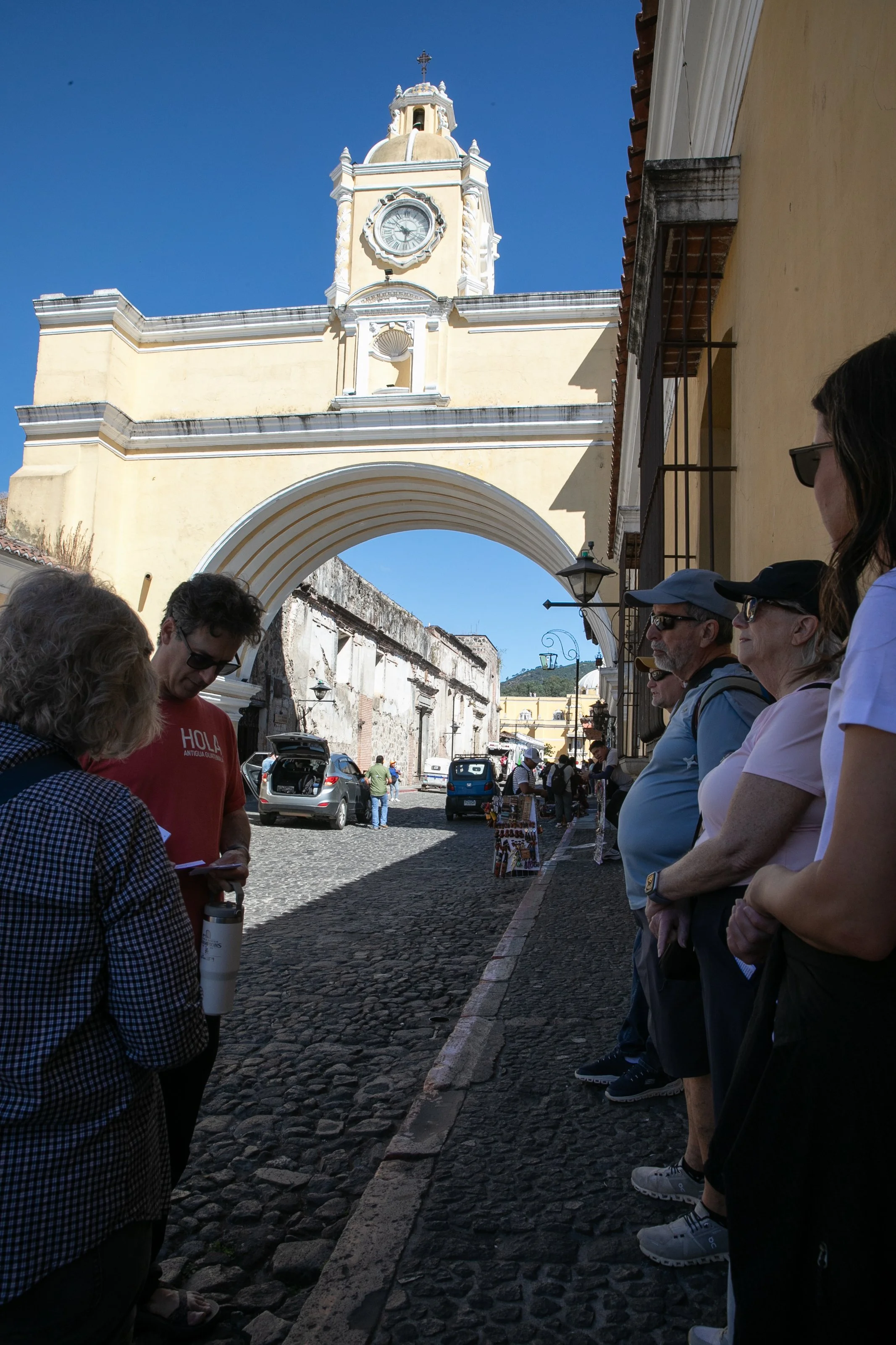 People standing in line outside a historic building with a clock tower, cobblestone street, and blue sky in the background, possibly in a Latin American country.