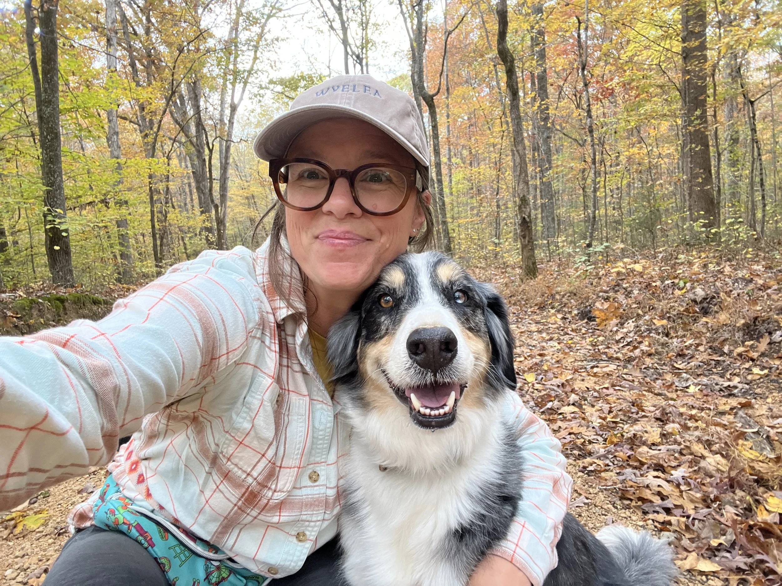 A woman wearing glasses and a beige cap taking a selfie in a forest with autumn-colored leaves while petting a happy Australian Shepherd dog.