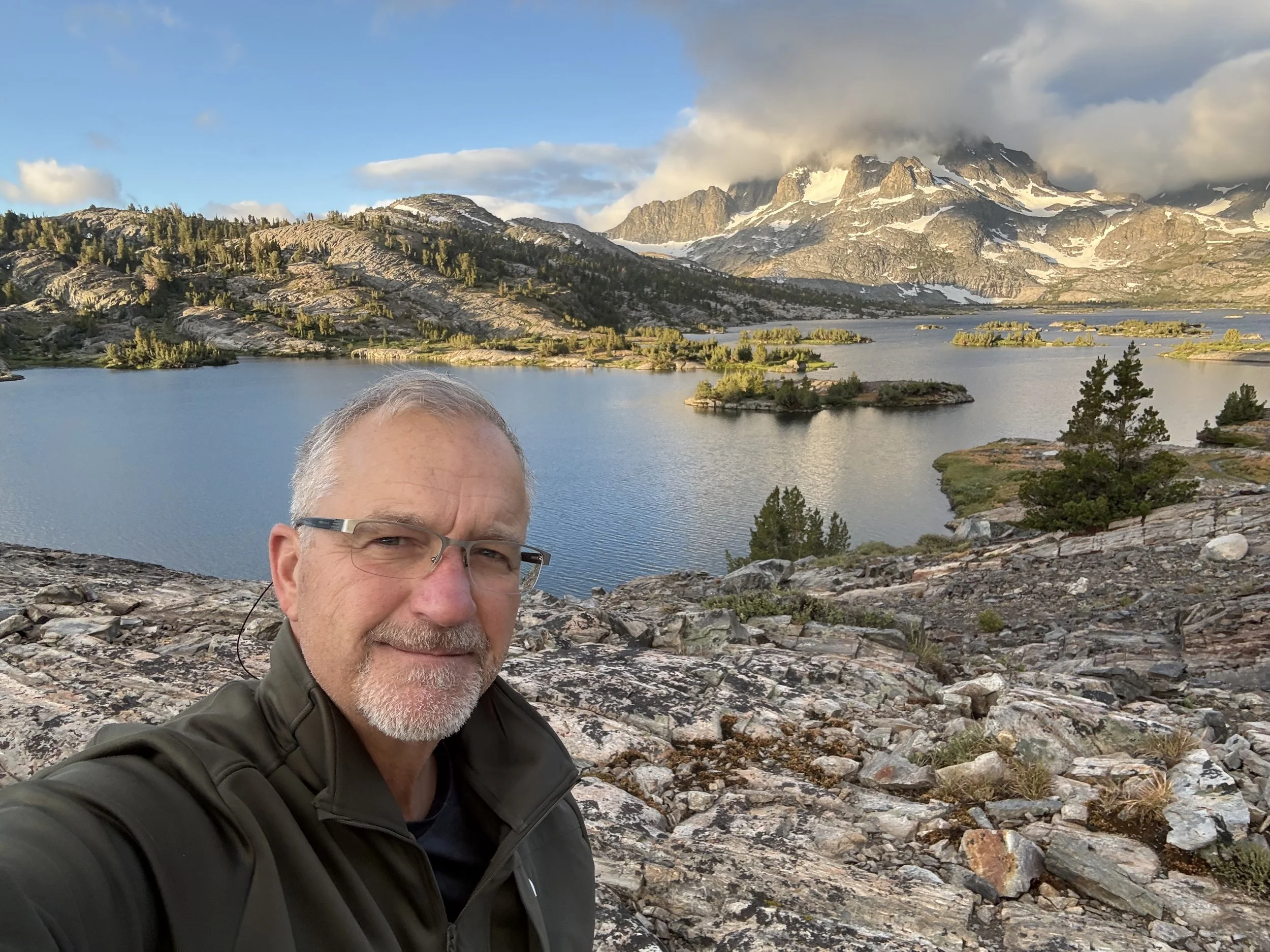A man with glasses and a gray beard taking a selfie with a mountain and lake landscape in the background, with rocky terrain in the foreground and snow-capped mountains partially covered by clouds.