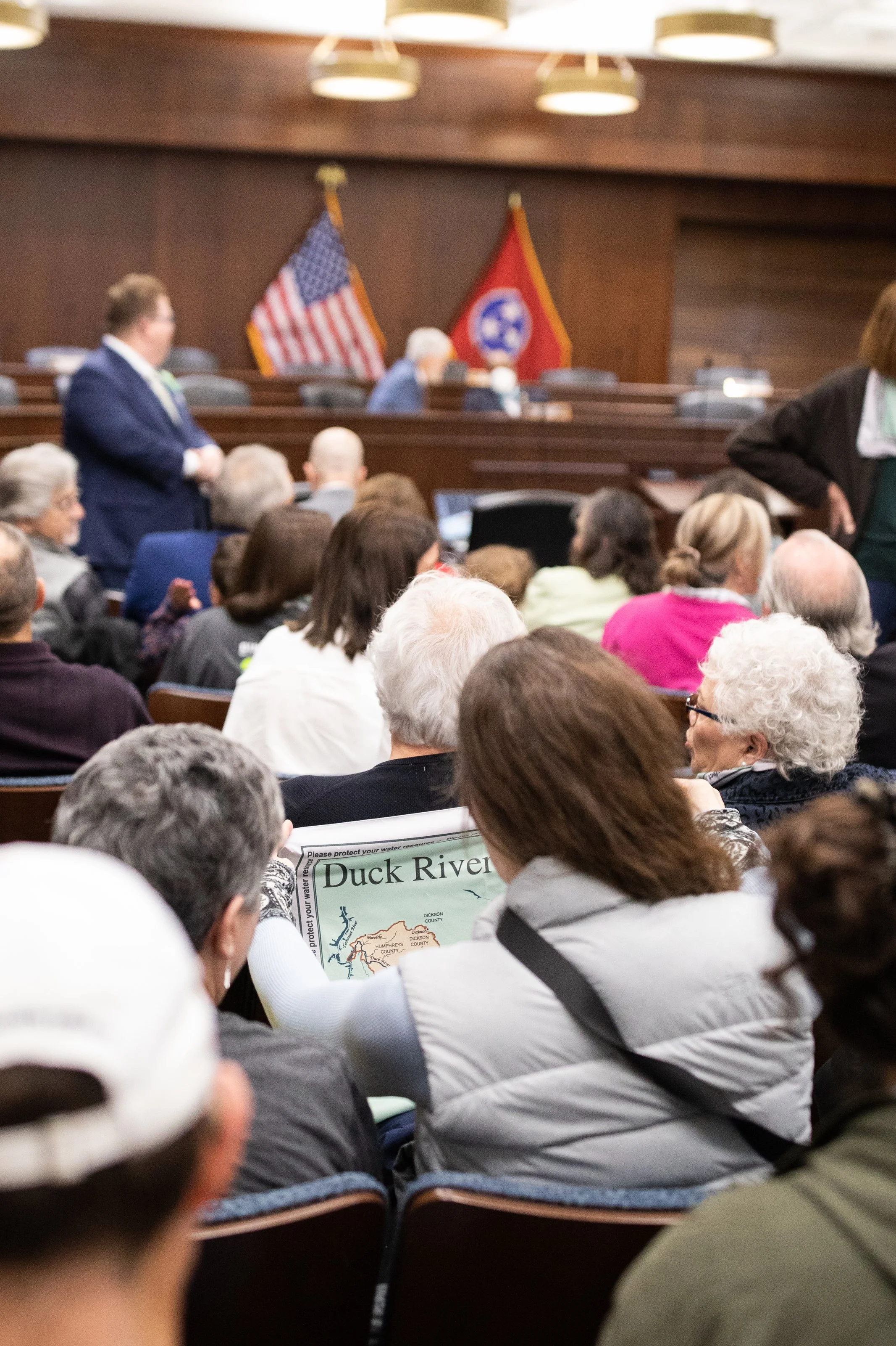 A political or town hall meeting with a diverse group of people seated and listening. Two flags, the United States flag and a Tennessee state flag, are displayed at the front of the room. Some attendees are taking notes or looking at documents.