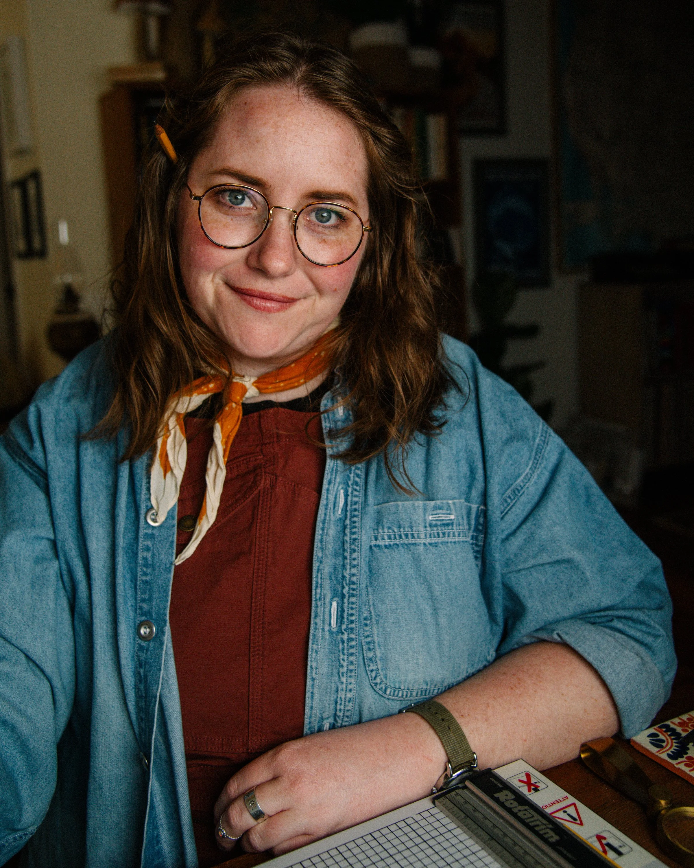 A woman with glasses, freckles, and wavy reddish-brown hair wearing a denim jacket, orange scarf, and a red top, sitting at a table with a graph paper notepad, a box cutter, and a board game with cards.