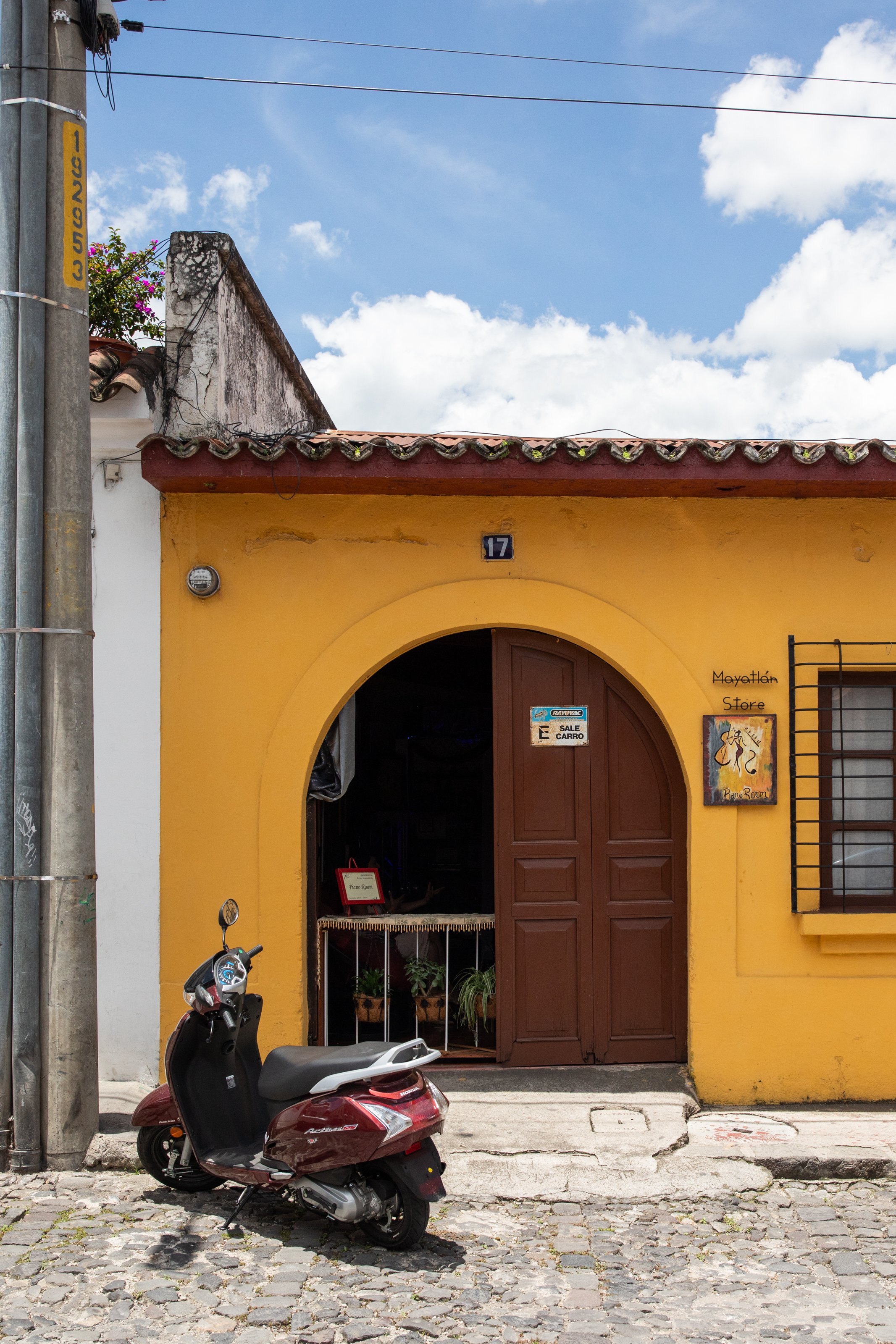 A red and black scooter parked in front of a yellow building with a brown wooden door and a sign indicating a piano room. The building has a small entrance with a potted plant inside, and a black metal decorative grille beside the door. There is a street with cobblestones in front of the building, and a scooter is parked nearby. The sky is partly cloudy.