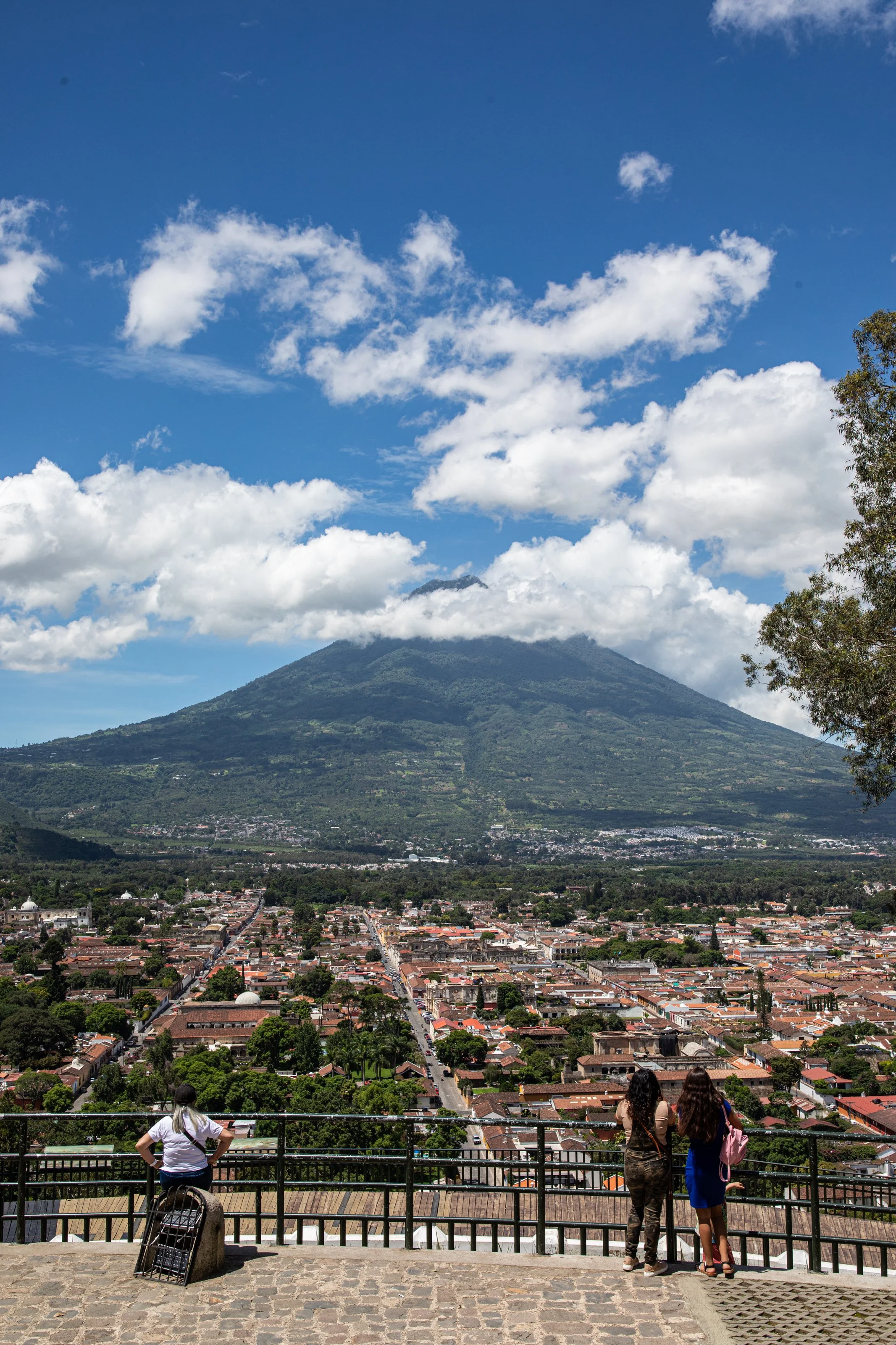 View of the city of Antigua Guatemala with a volcano in the background, seen from the Mirador de la Cruz observation point, with three women looking at the scenic view.