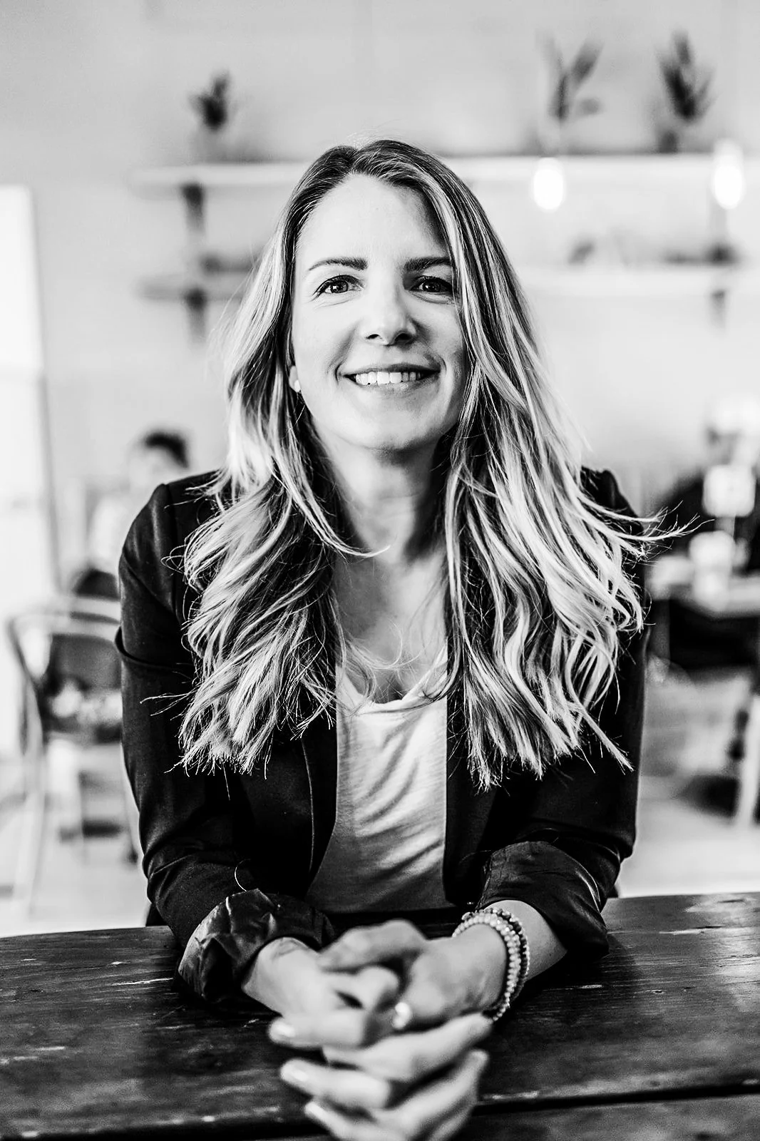 A woman with long wavy hair smiling, sitting at a table in a cafe. The photo is black and white.