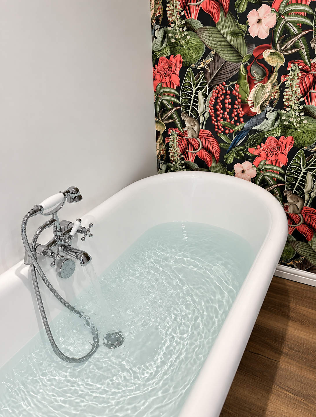 A white vintage bathtub filled with water, with chrome fixtures, positioned next to a wall with botanical-themed wallpaper featuring tropical plants, flowers, a monkey, and a bird, and a wooden floor.