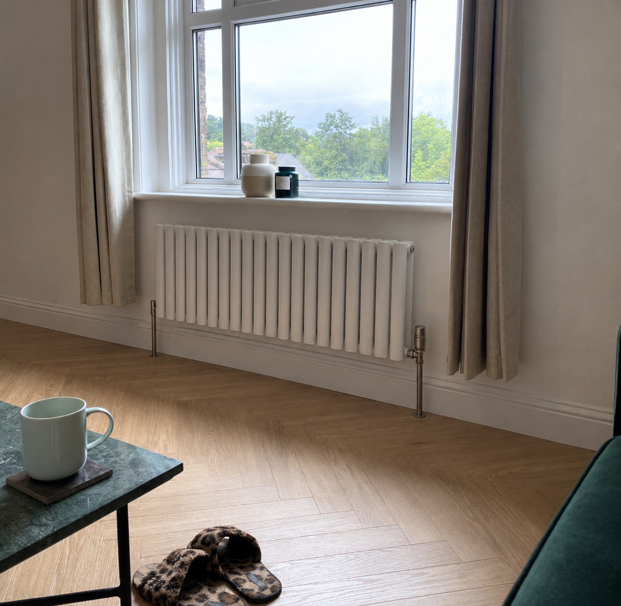 A room with a window, beige curtains, a white radiator, and a wooden floor. On the window sill are two vases and a candle. In the foreground, a green table with a mug and a pair of leopard print slippers on the floor.