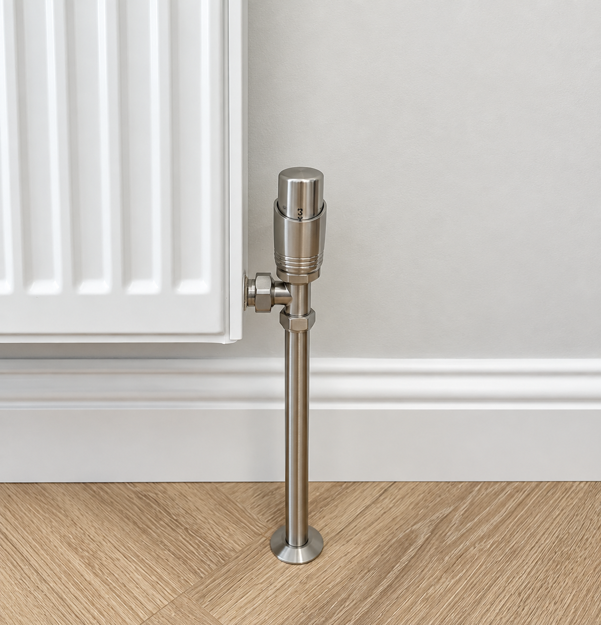 Close-up of a metallic radiator valve pipe connected to a radiator in a room with wooden flooring and light-colored walls.