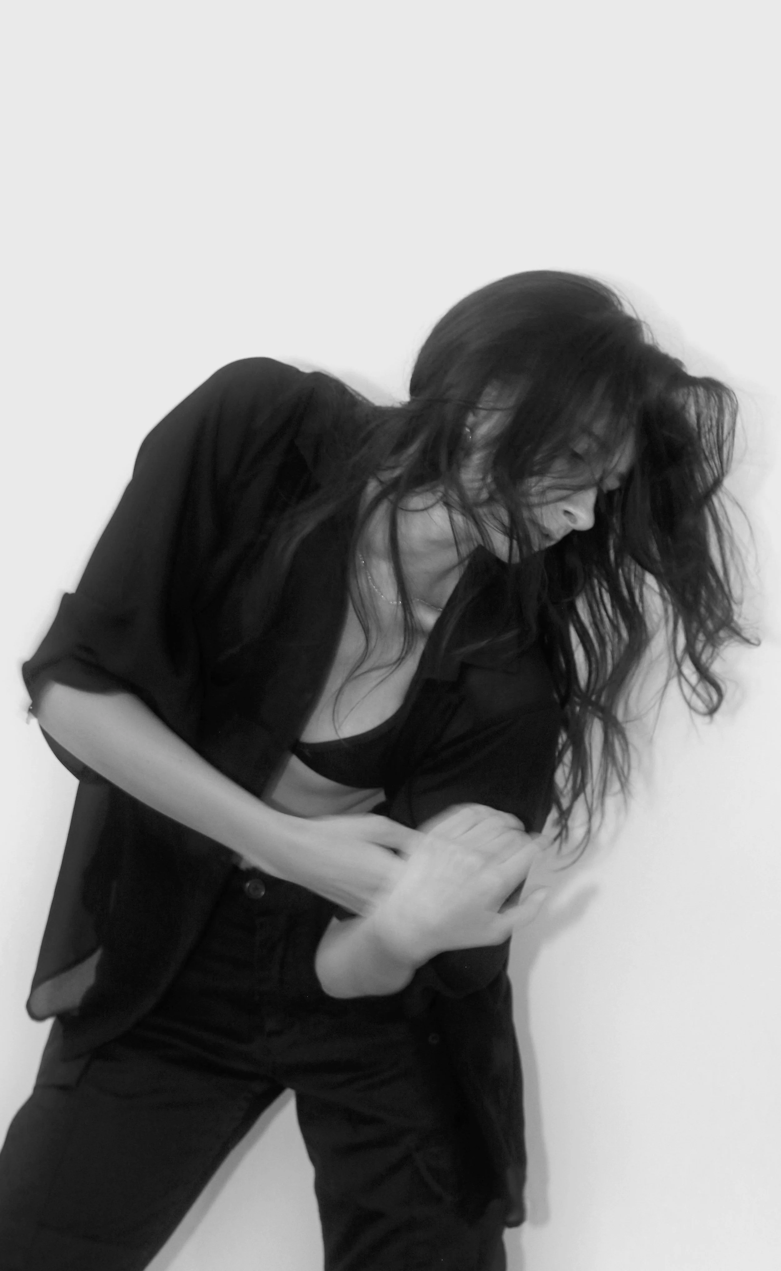 Black and white photo of a woman with long, wavy hair looking down at her phone, wearing a loose black blazer and pants, standing against a plain white wall.