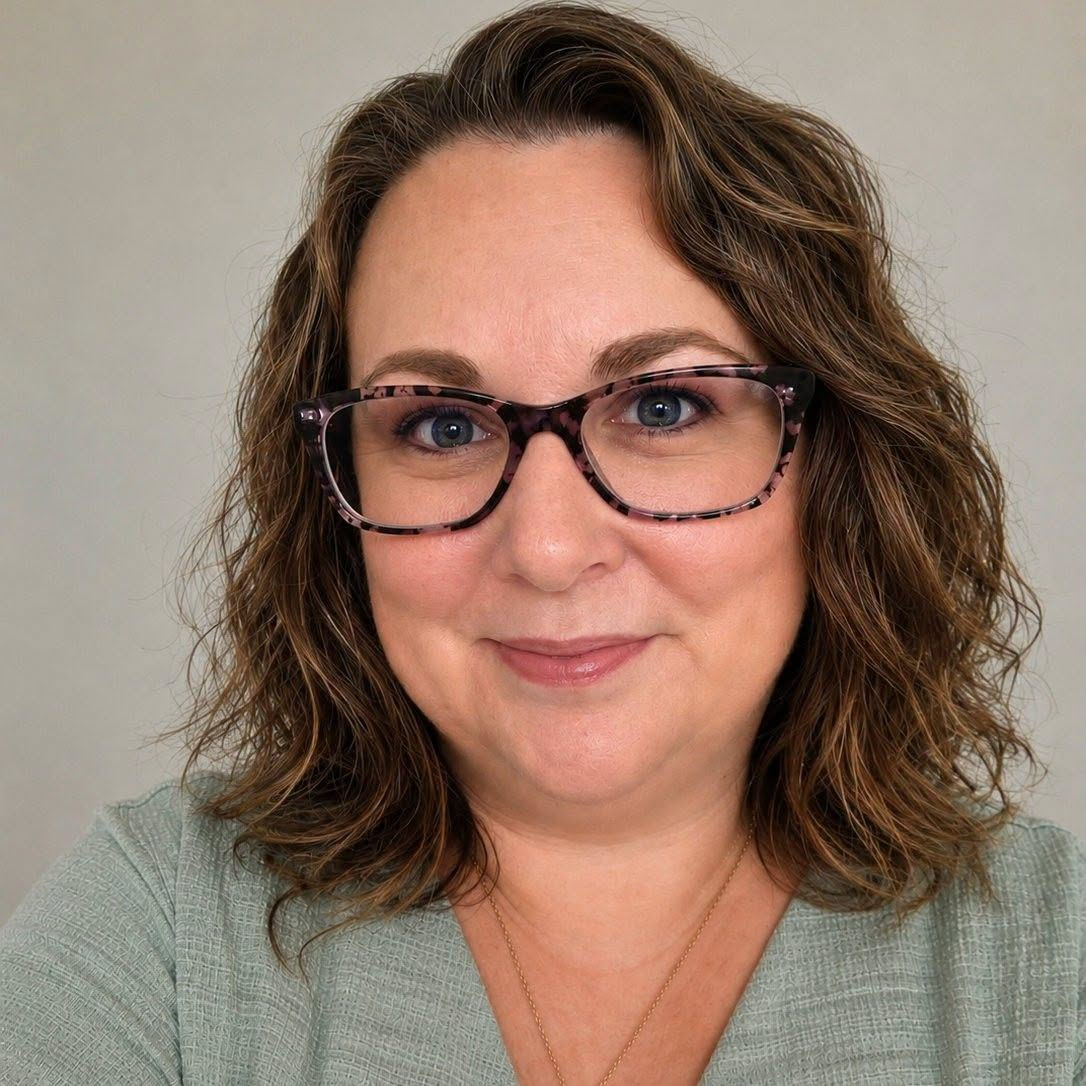 A woman with shoulder-length curly brown hair wearing glasses with a tortoiseshell pattern and a light green top, smiling against a neutral background.