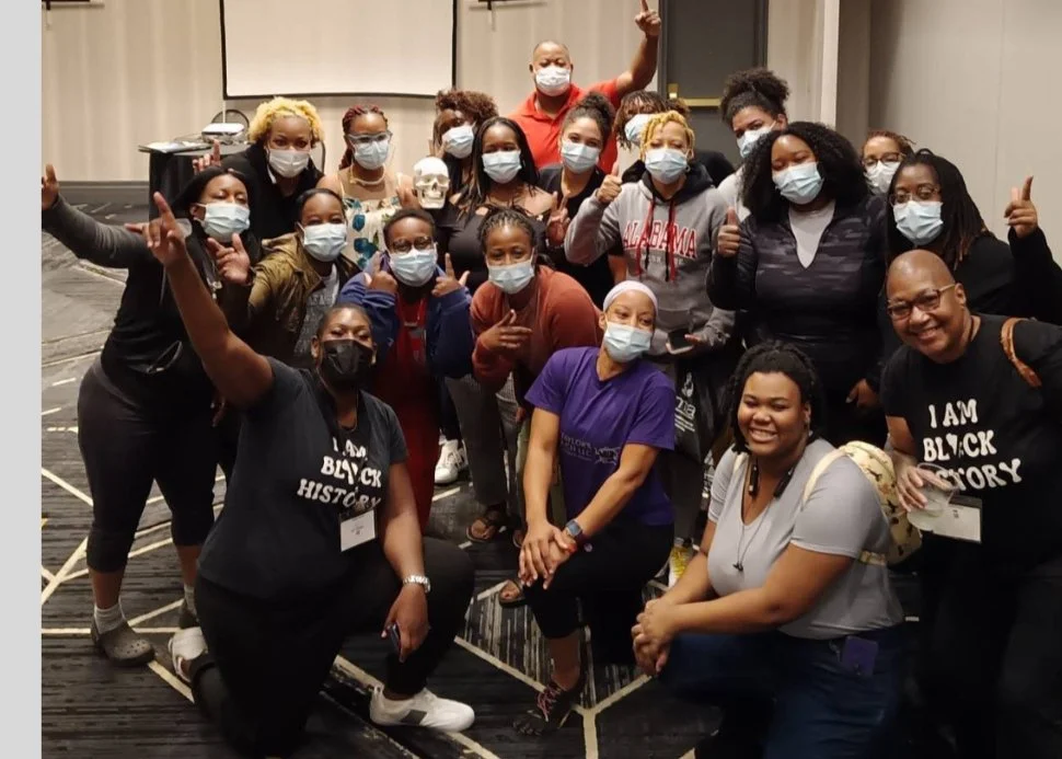 A diverse group of people, mostly women, gathered together wearing masks, some holding a skull, in an indoor setting, celebrating or participating in an event related to Black history or culture.