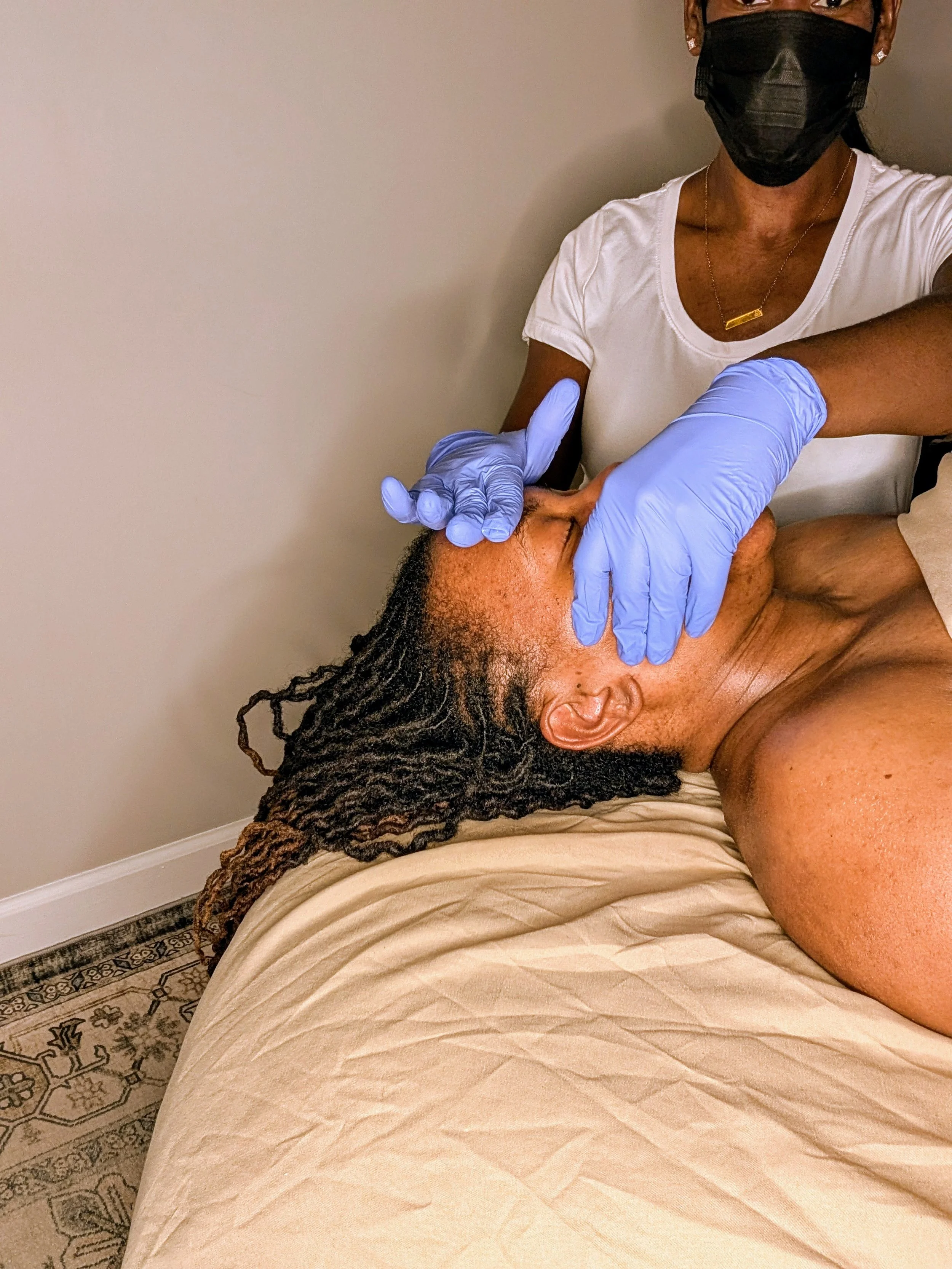 A woman lying on a massage table receiving intra-oral tmj massage from AU massage therapist wearing blue gloves and a face mask.