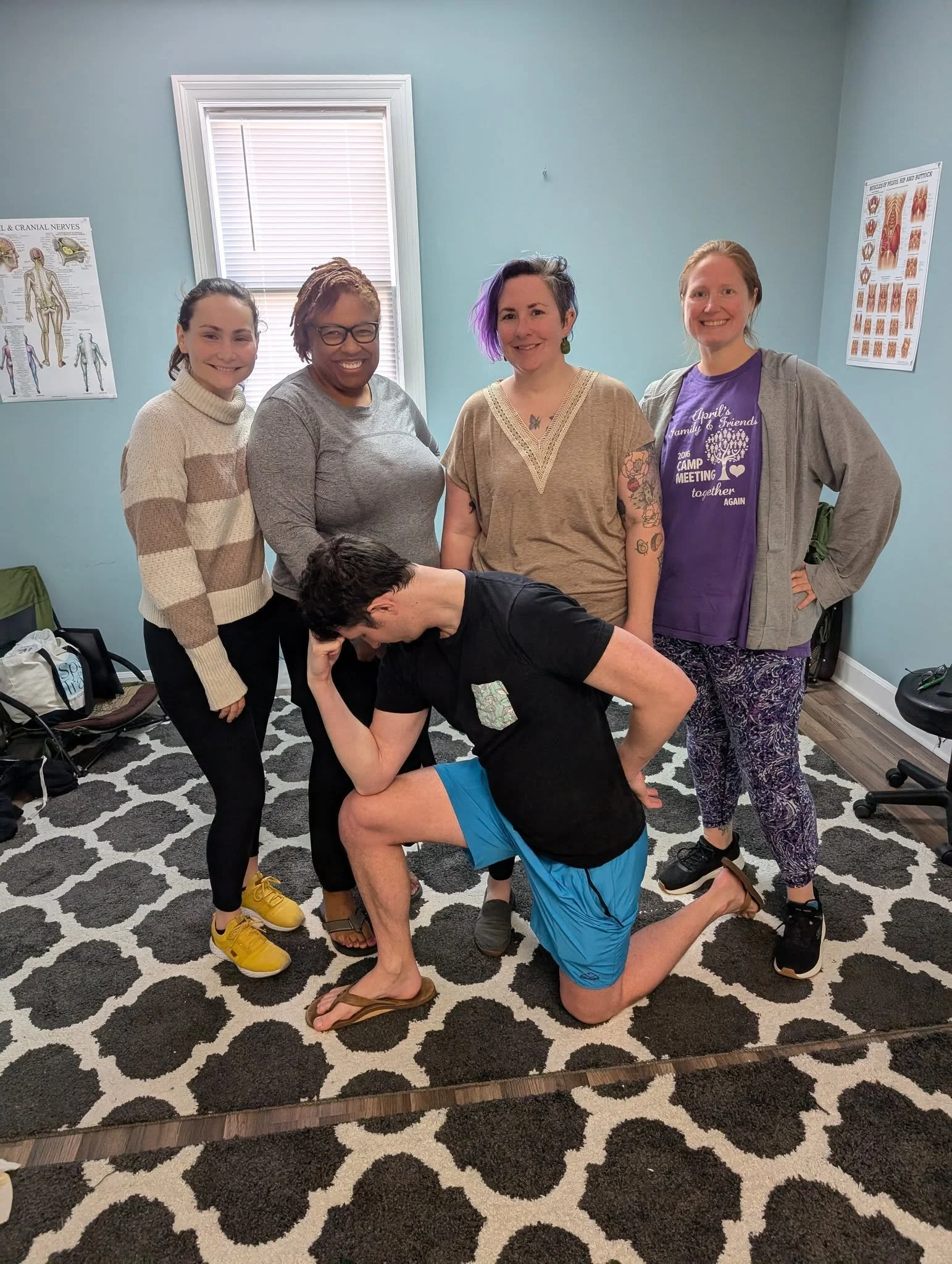 Four women and one man in a room with blue walls and a black and white patterned carpet. The women are standing and smiling, with one woman in the foreground in a purple t-shirt that reads 'April's Family & Friends 2016 Camp Meeting together again,' and another woman with purple hair. The man is holding a plank and kneeling in front of them, with his head bent down. There are posters on the walls and belongings on the floor.