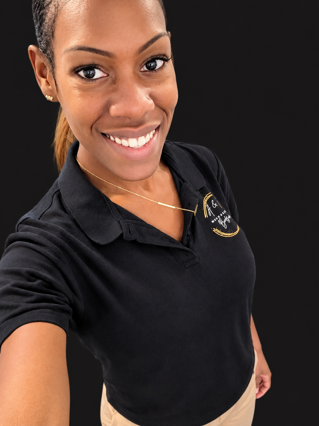 A smiling woman taking a selfie against a black background. She has dark hair tied back, wearing earrings, a gold necklace, a black polo shirt with gold embroidery, and beige pants.