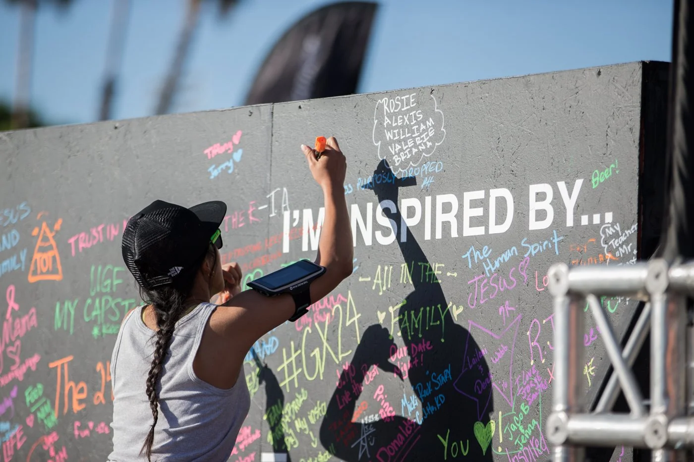 A girl with a braid, wearing a black cap and white tank top, writes with multicolored chalk on a large gray wall. The wall is decorated with various messages, signatures, and drawings in different colors, including the phrase "I'm inspired by..." in large white letters. The girl’s shadow is cast onto the wall, and she has a smartwatch on her left arm.