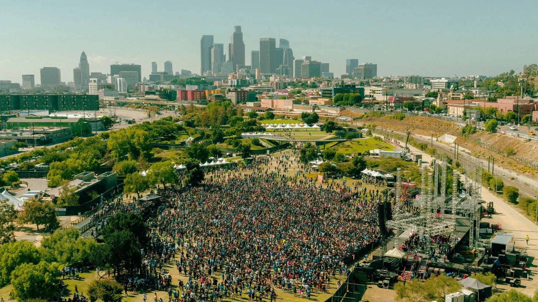 Overhead view of a large outdoor concert in a park with a dense crowd, tents, and stage equipment, with a city skyline in the background.