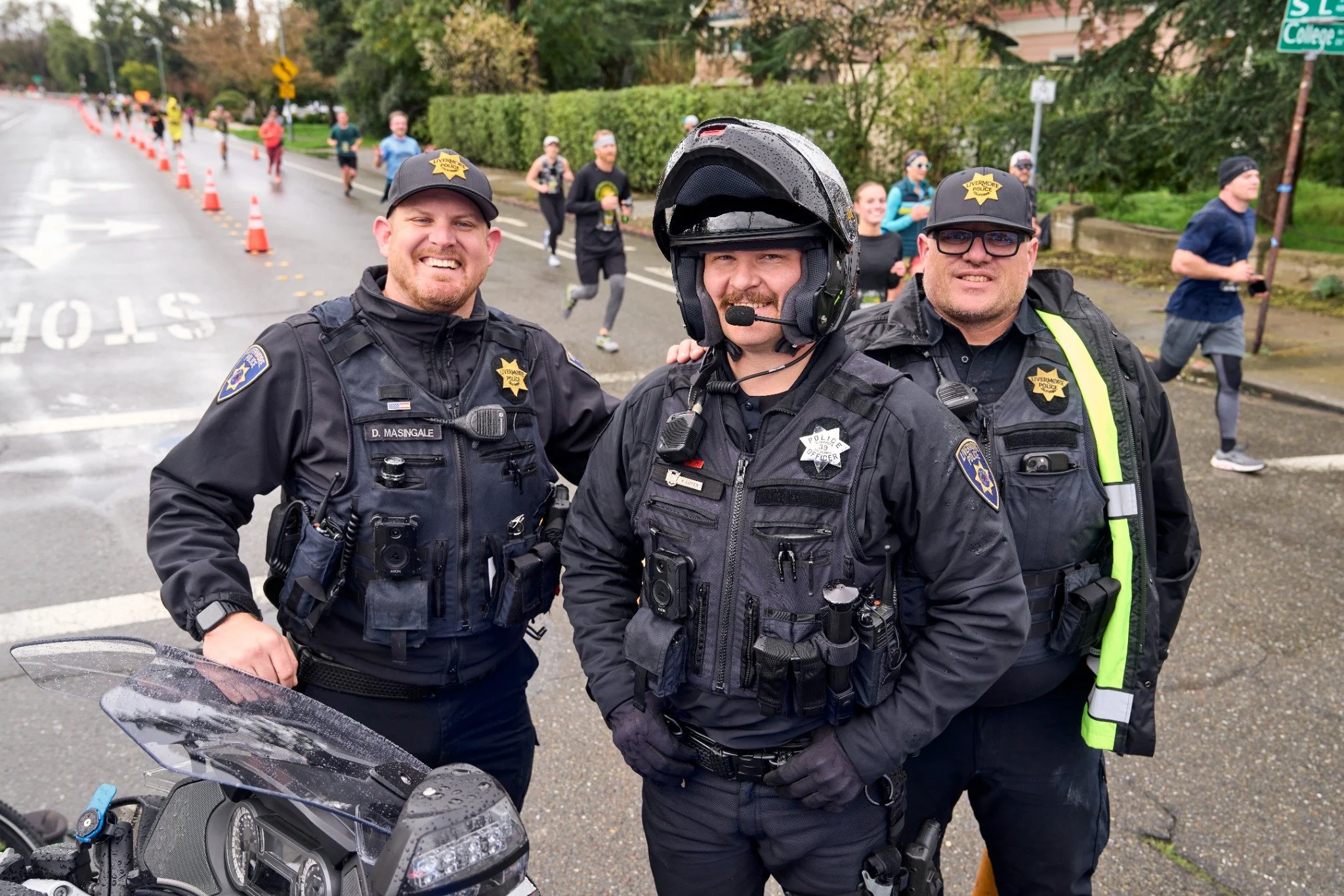 Three police officers standing in front of a group of runners in a race. Two officers wear black uniforms with police badges, one of them is wearing a helmet, and all are smiling. The runners are in the background on a wet street with orange cones and greenery.
