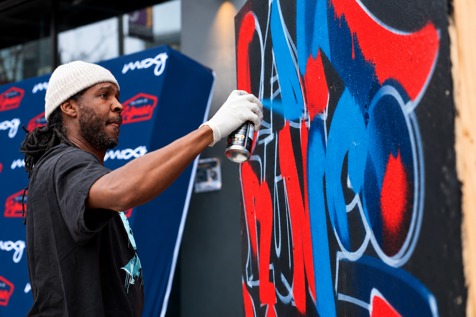 A man with dreadlocks, wearing a beige knit hat, spray-painting graffiti in red, blue, and black on a large black wall outdoors.