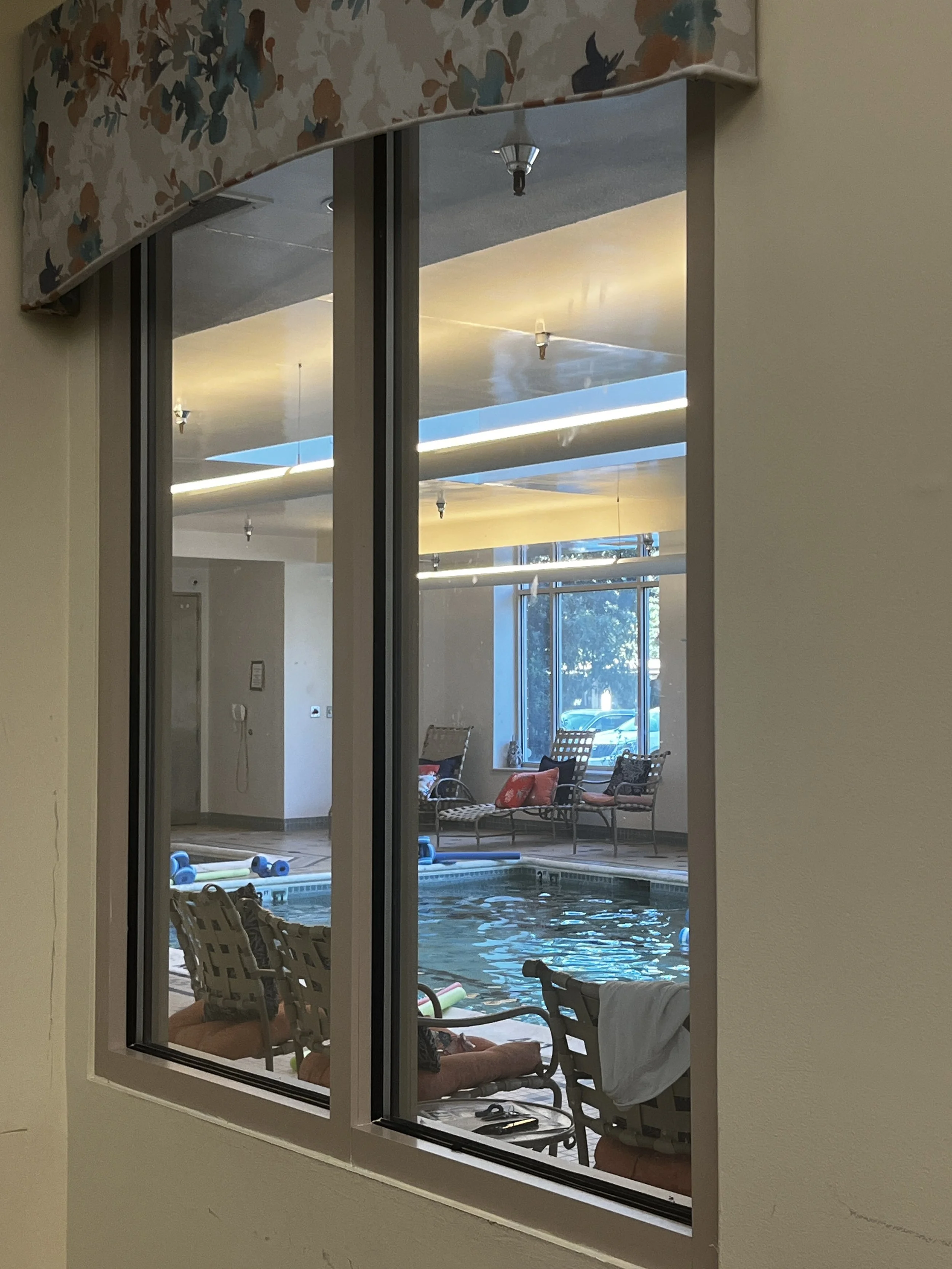 View through a window into an indoor pool area with chairs, a person relaxing, pool floats, and fitness equipment, illuminated by natural and artificial light.