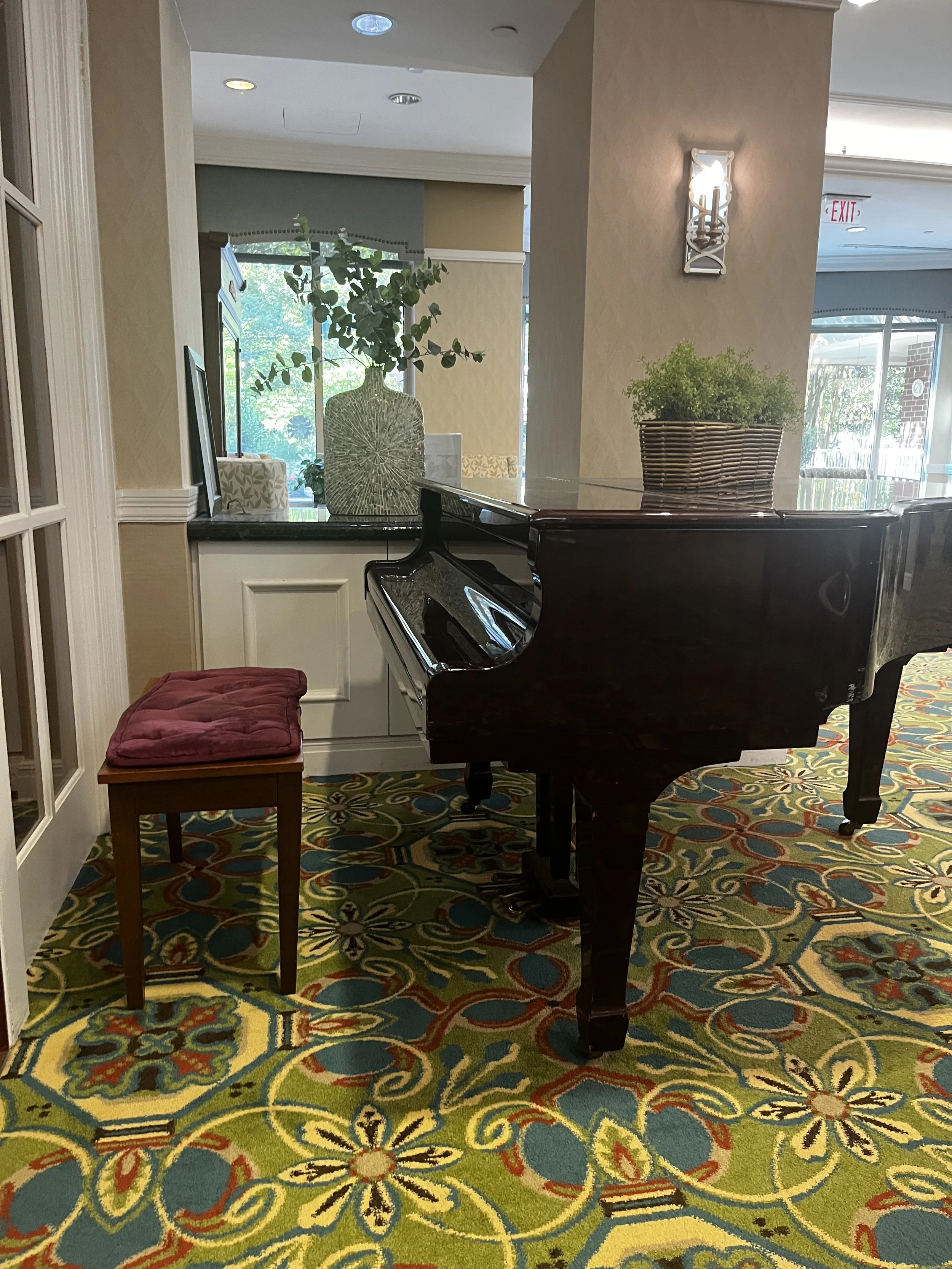 A grand piano with a dark wood finish, a small wooden stool with a red cushioned seat in front, and a decorative floral-patterned carpet under a large window in a cozy room.