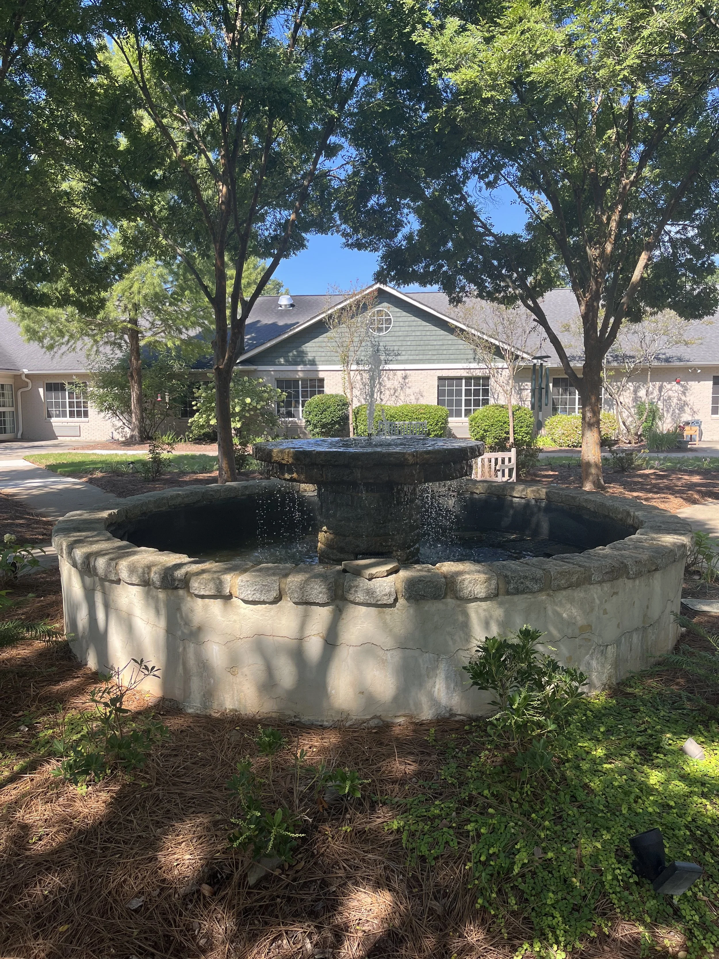 A stone fountain with water in a garden surrounded by trees and shrubs, with a house in the background.