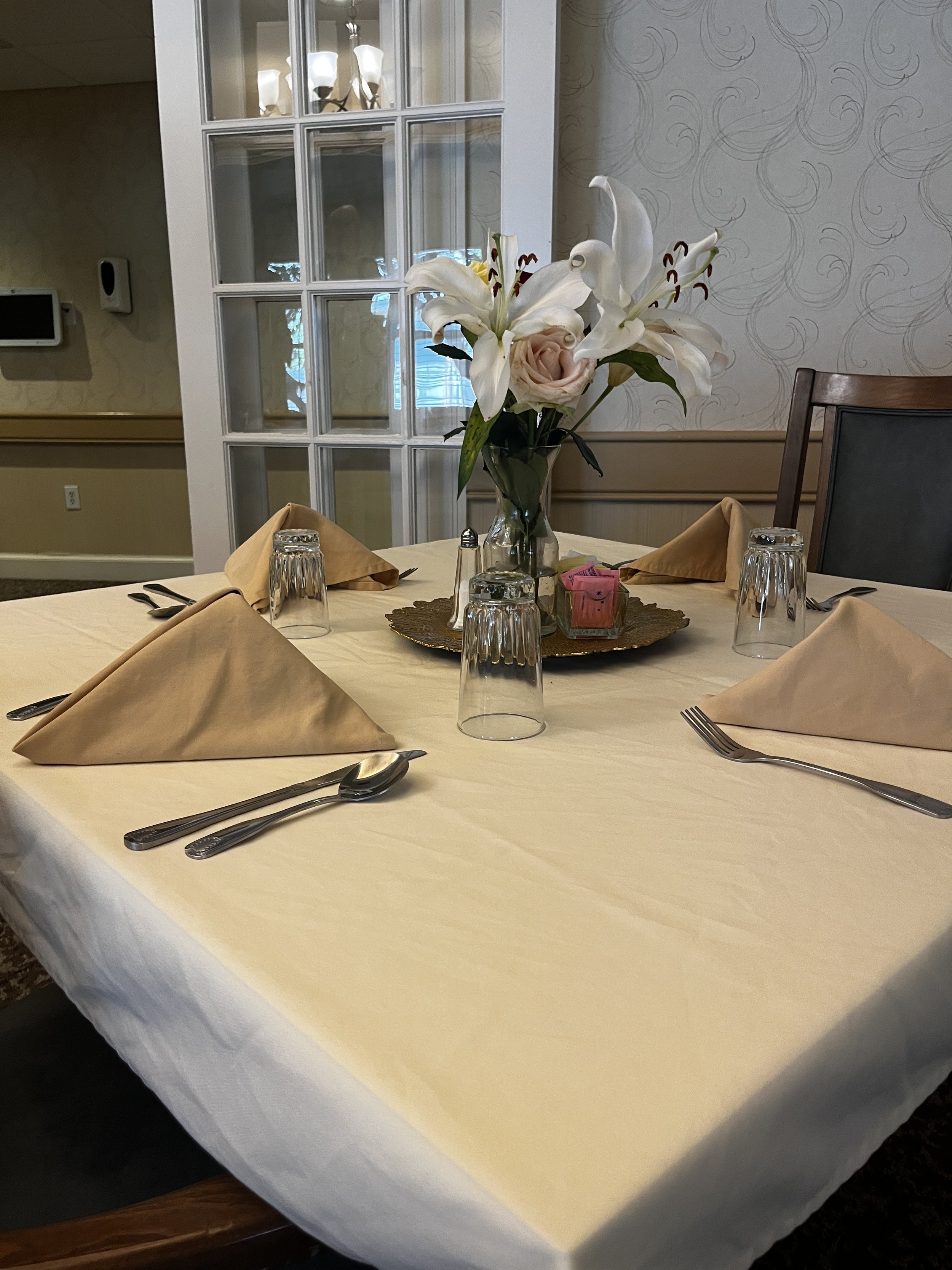 Elegant dining table set with beige napkins, glasses upside down, silverware, and a centerpiece of white lilies and a pink rose in a glass vase, on a cream tablecloth.