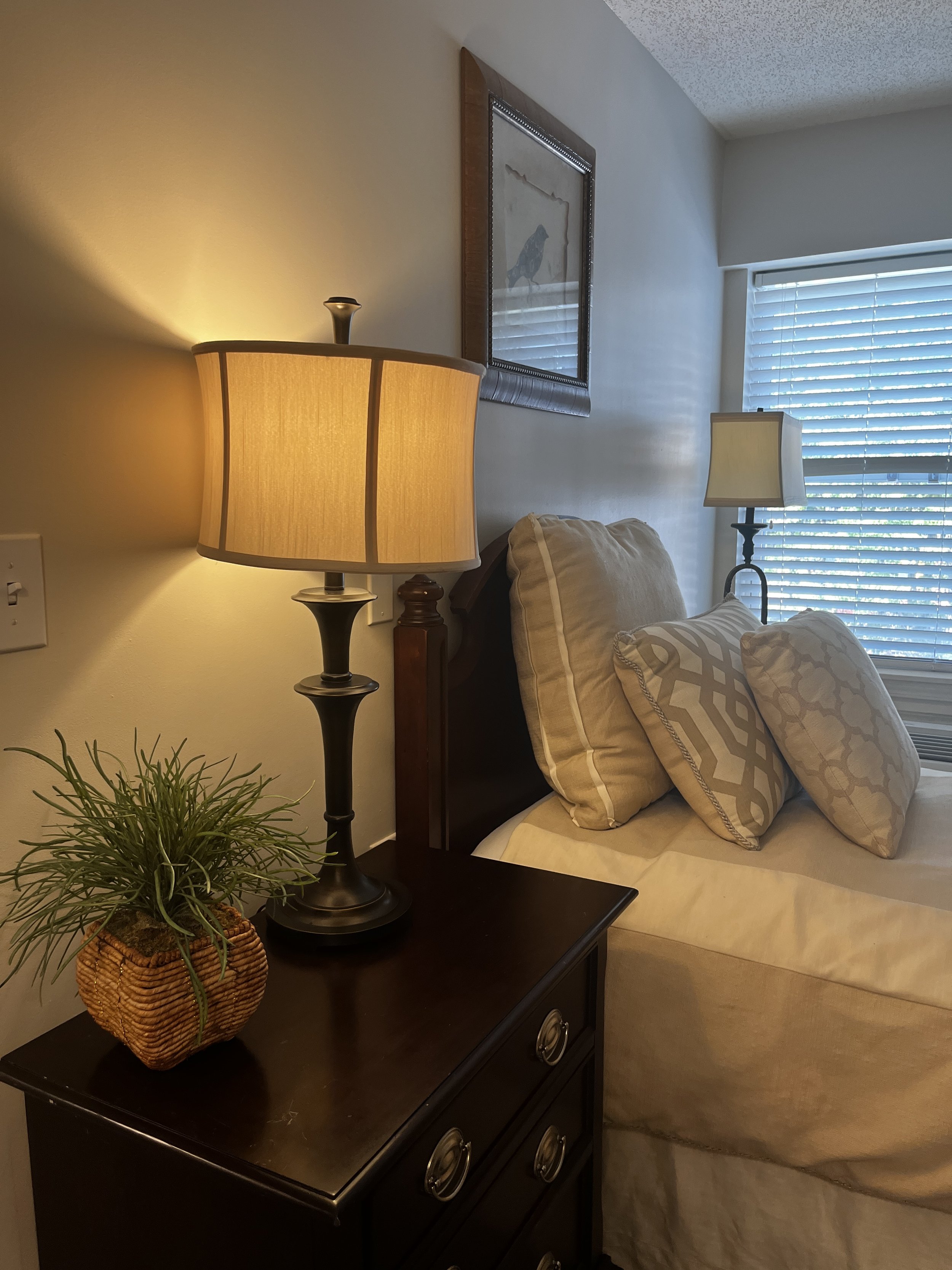 A bedroom corner with a dark wood nightstand, a potted plant in a wicker basket, two table lamps, beige pillows on the bed, a framed painting of a bird on the wall, and closed white blinds covering a window.