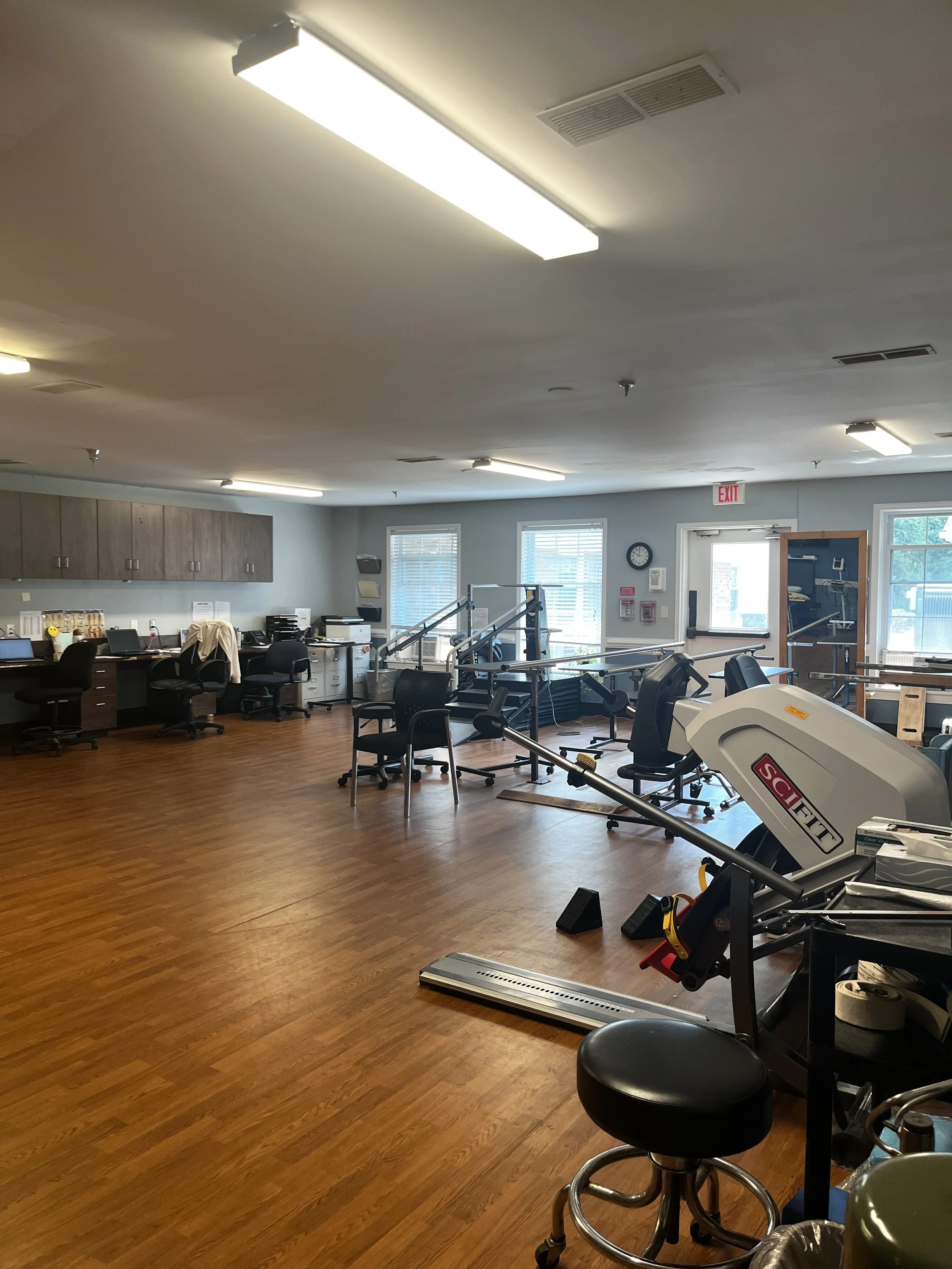 A spacious physical therapy or rehabilitation clinic with various exercise equipment, chairs, and a wooden floor, illuminated by overhead fluorescent lights, with windows on the far wall and an exit sign above a door.
