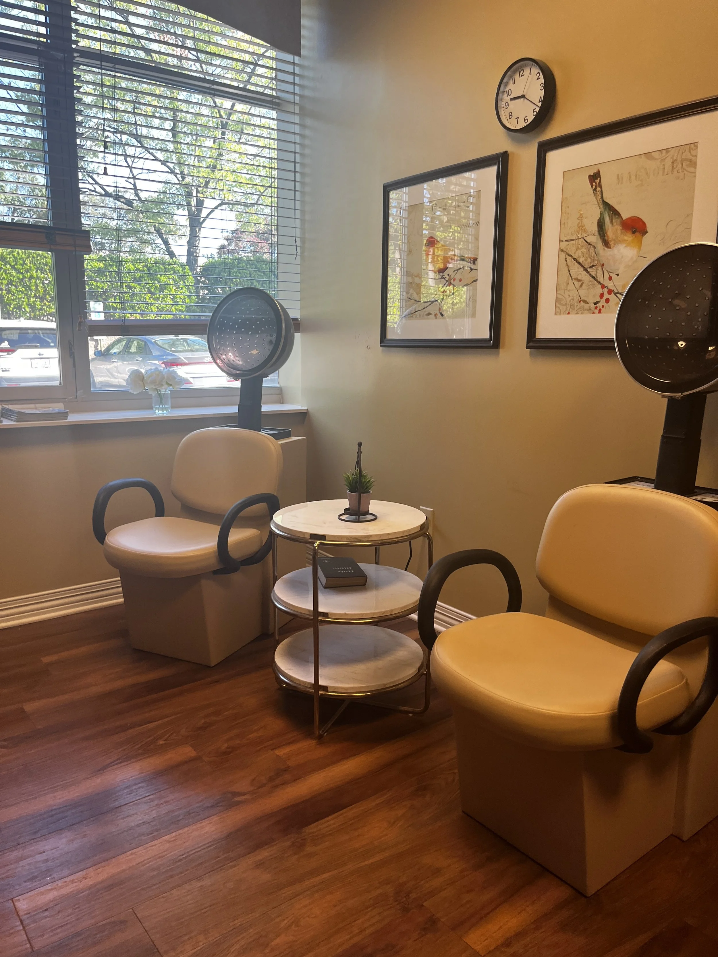 A cozy indoor seating area with yellow chairs, a small white round table, and artwork of birds on the wall. There is a window with blinds letting in natural light, and a clock showing 3:24. The floor is wooden.
