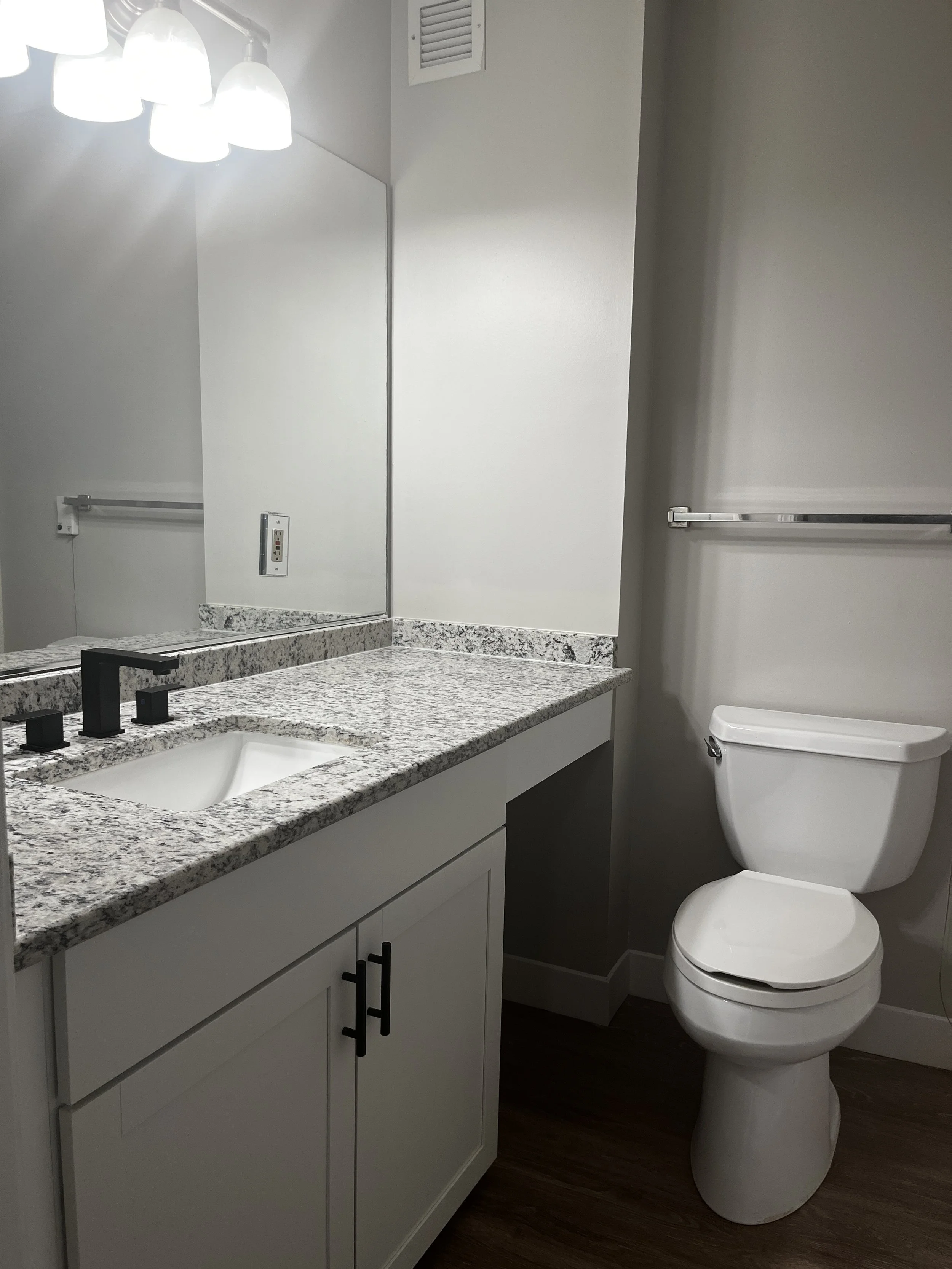 A bathroom with a granite countertop sink, mirrored wall, electrical outlet, towel rack, and a toilet.